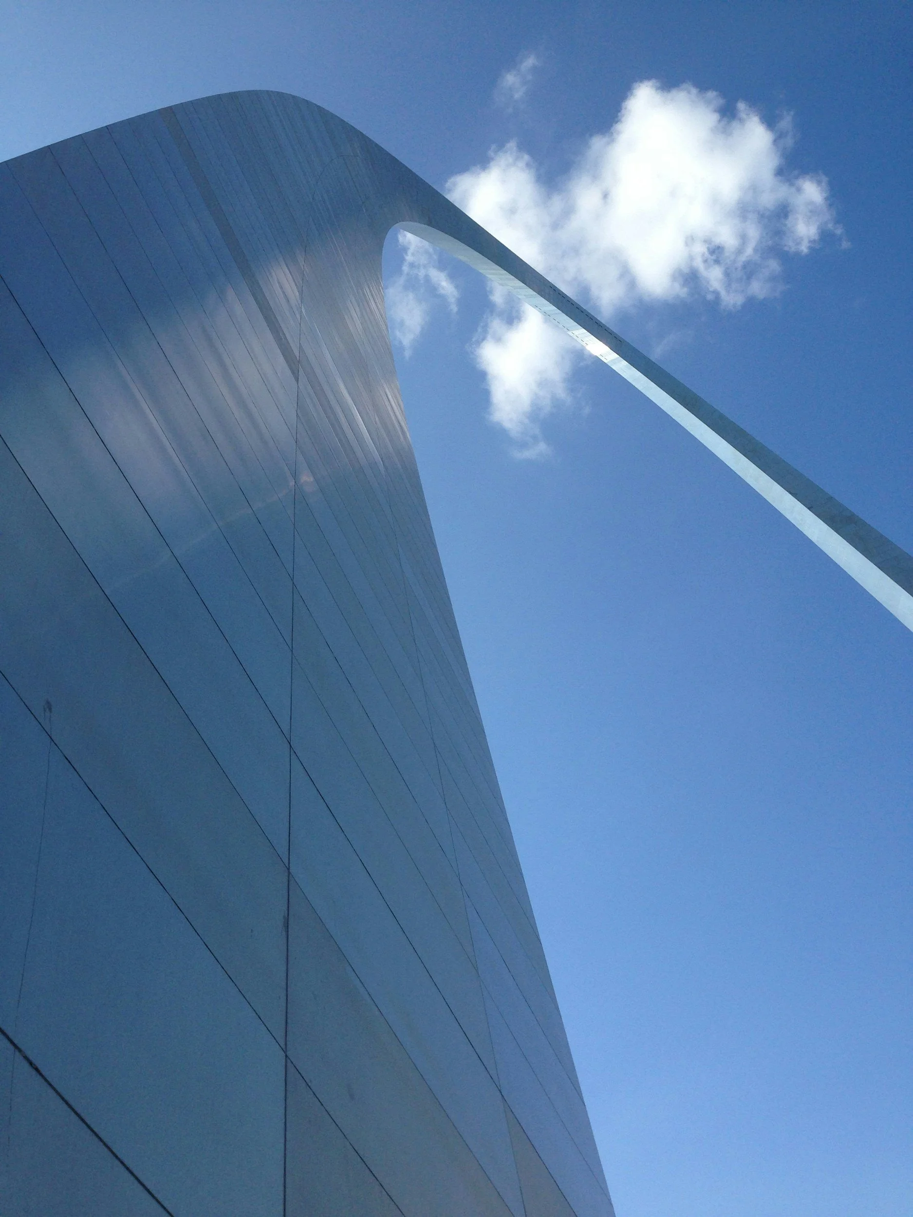 Looking up at the St. Louis Arch: a smooth, shiny surface, set against a bright blue sky with scattered white clouds.