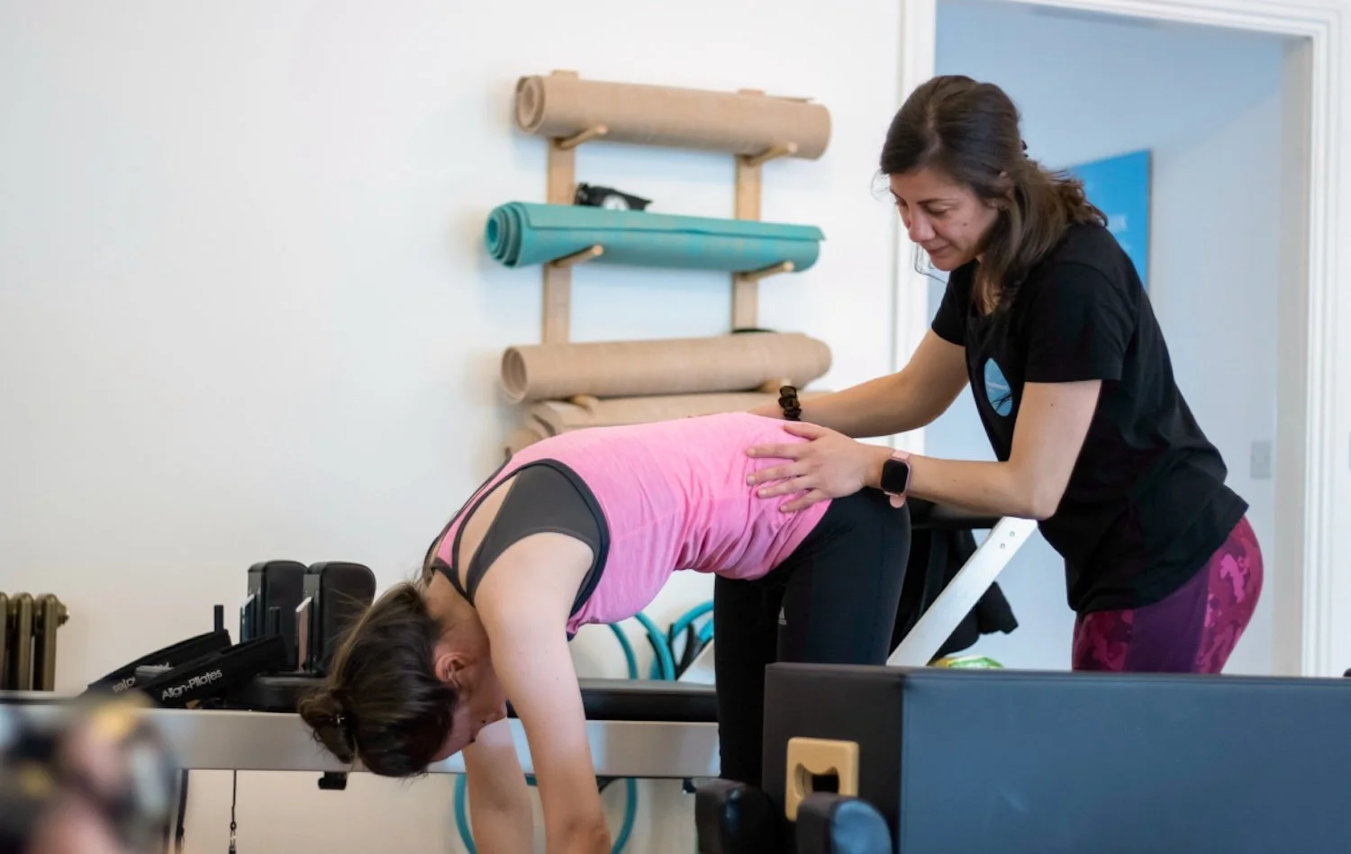 A female physiotherapist assisting a woman in a pink tank top with a pilates exercise on a reformer machine in a pilates studio.