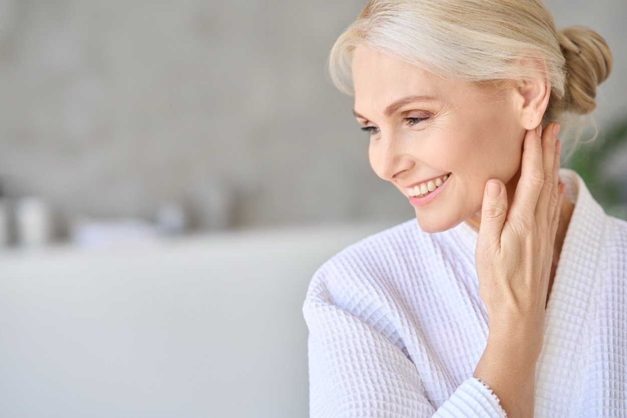 A smiling female with light grey hair touching her neck in a bright room.