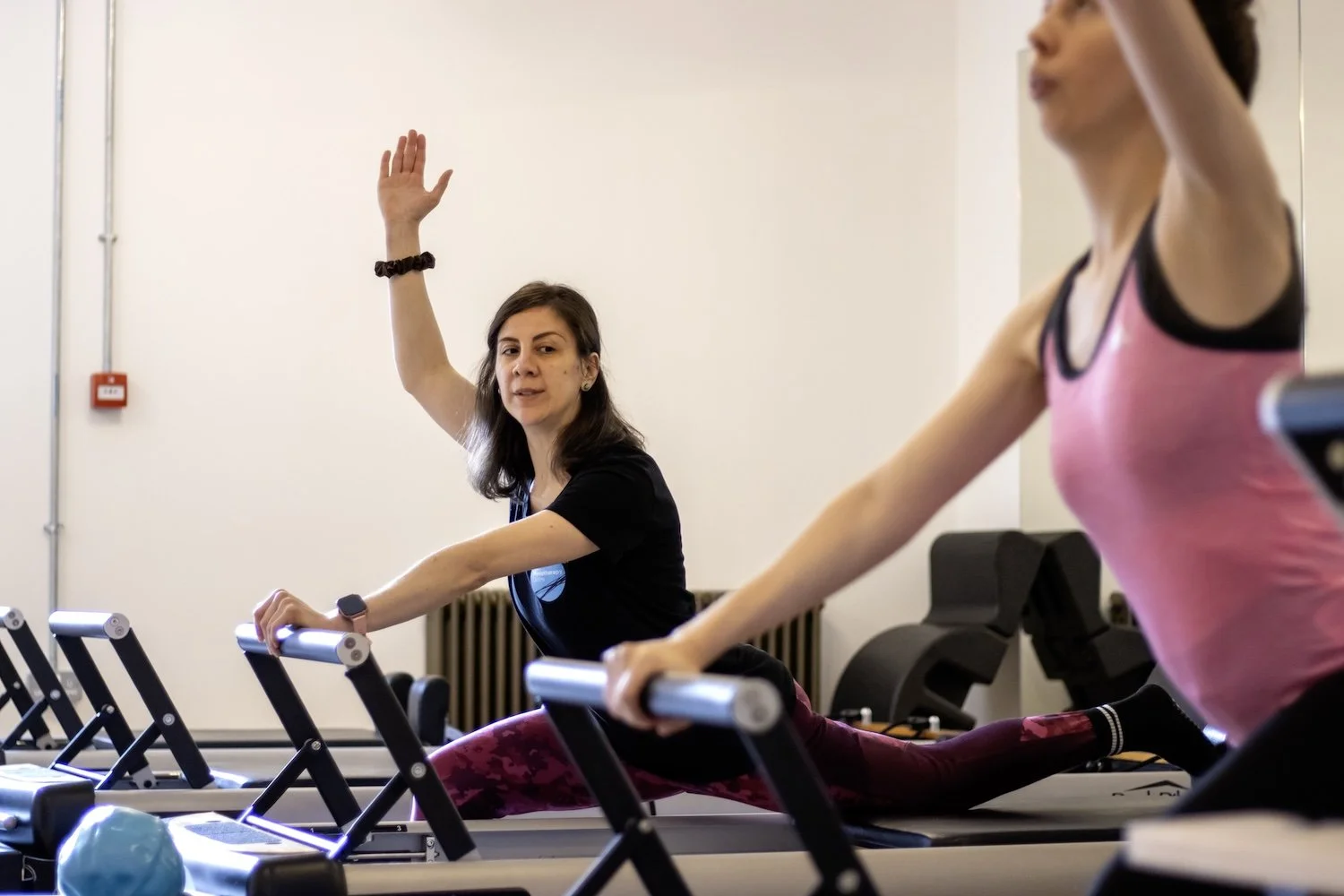 Female pilates instructor with one arm in the air, demonstrating an exercise to a client on reformer pilates equipment.
