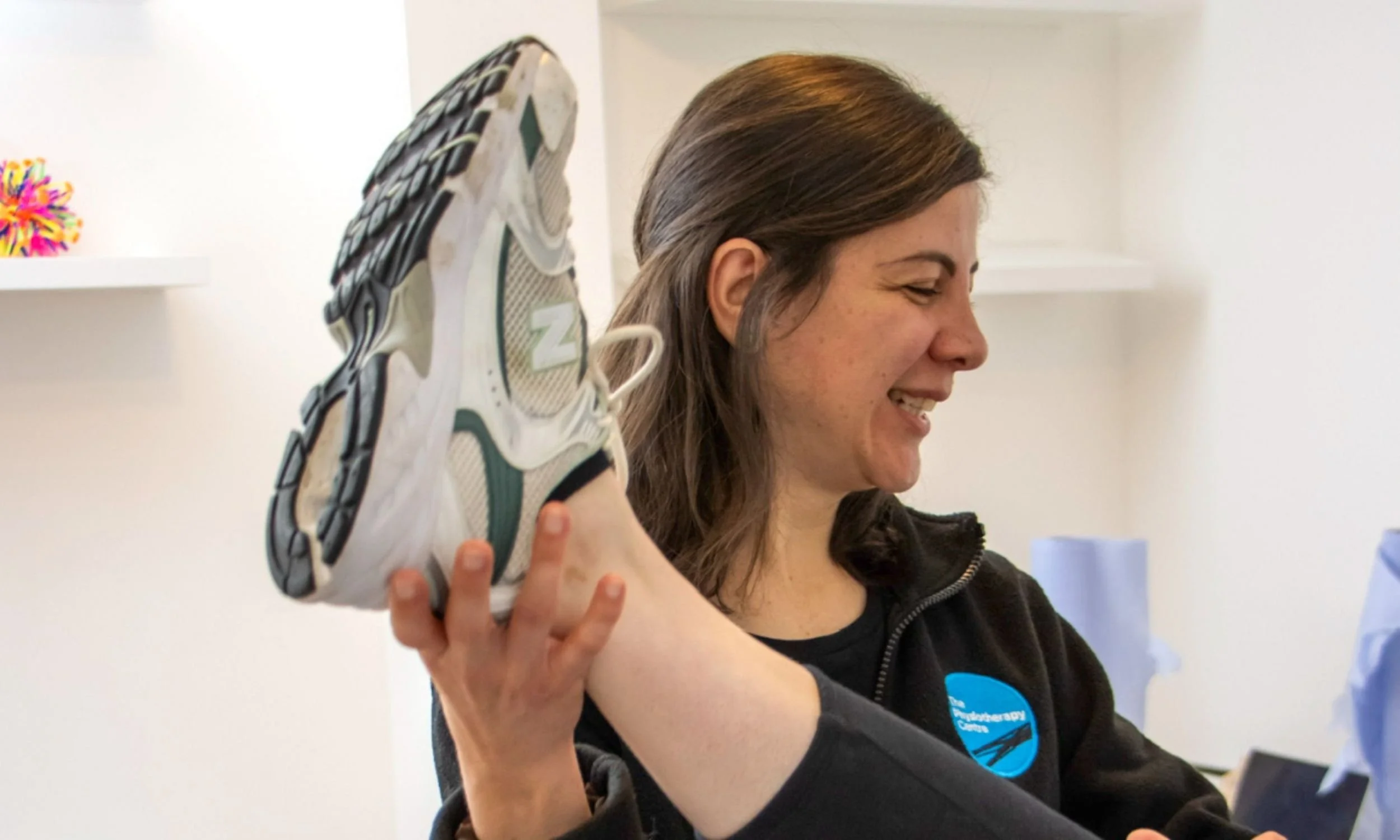 Female physiotherapist with long brown hair smiling as she holds a running shoe near her shoulder in a stretch.