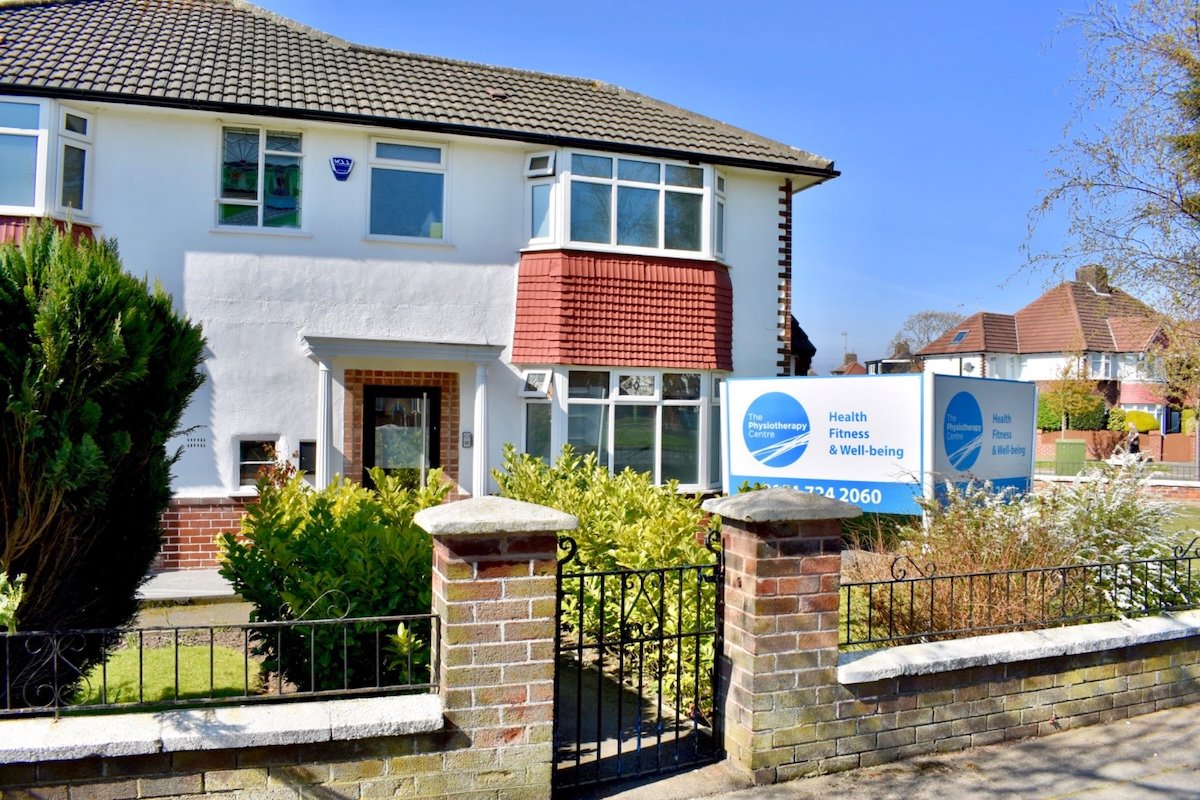 A physiotherapy centre with a sign in front that reads 'The Physiotherapy Centre, Health, Fitness & Well-being.' The house has a white exterior with a brick entryway and a garden with bushes and trees. The sky is clear and blue.