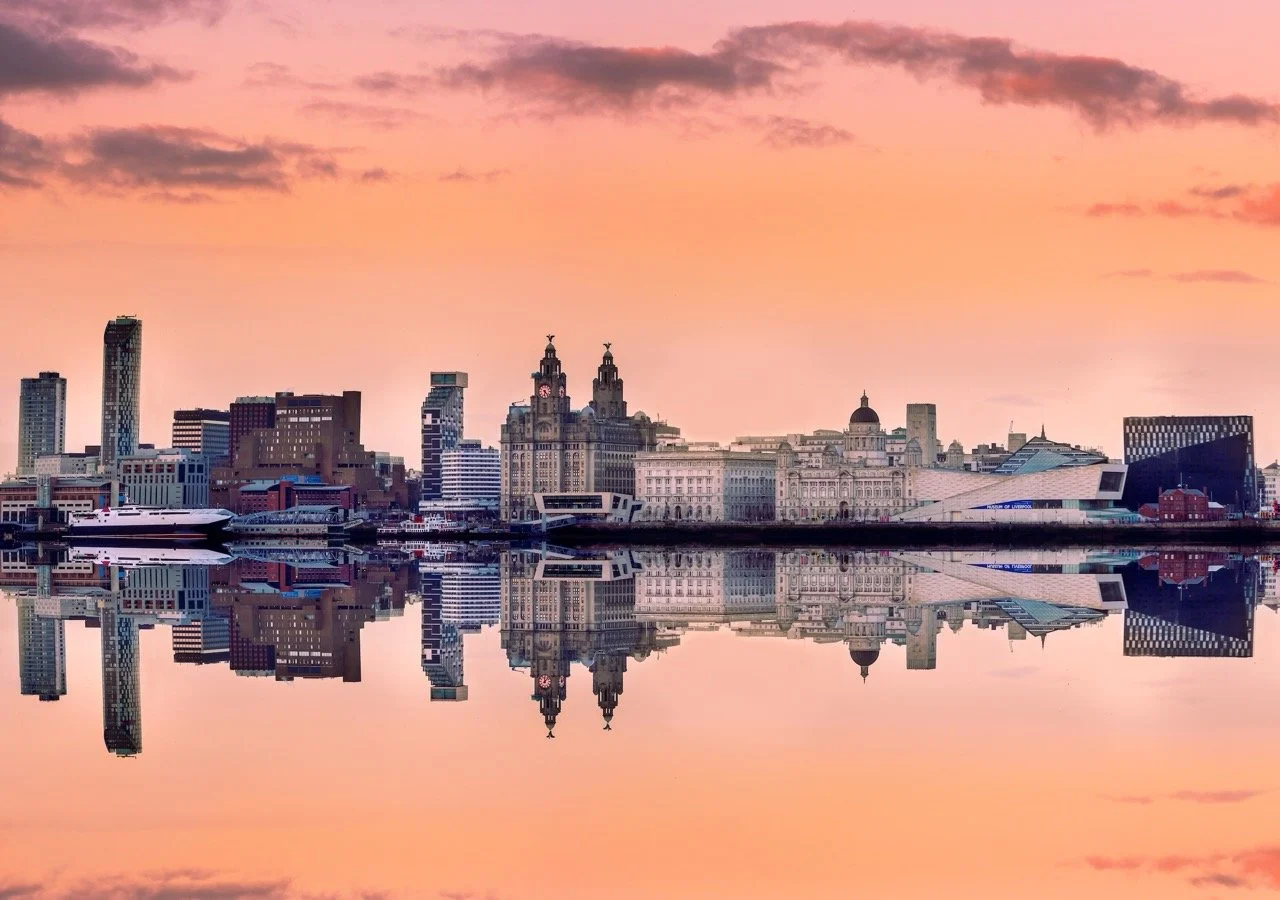 Liverpool city skyline at sunset with buildings reflected in water.