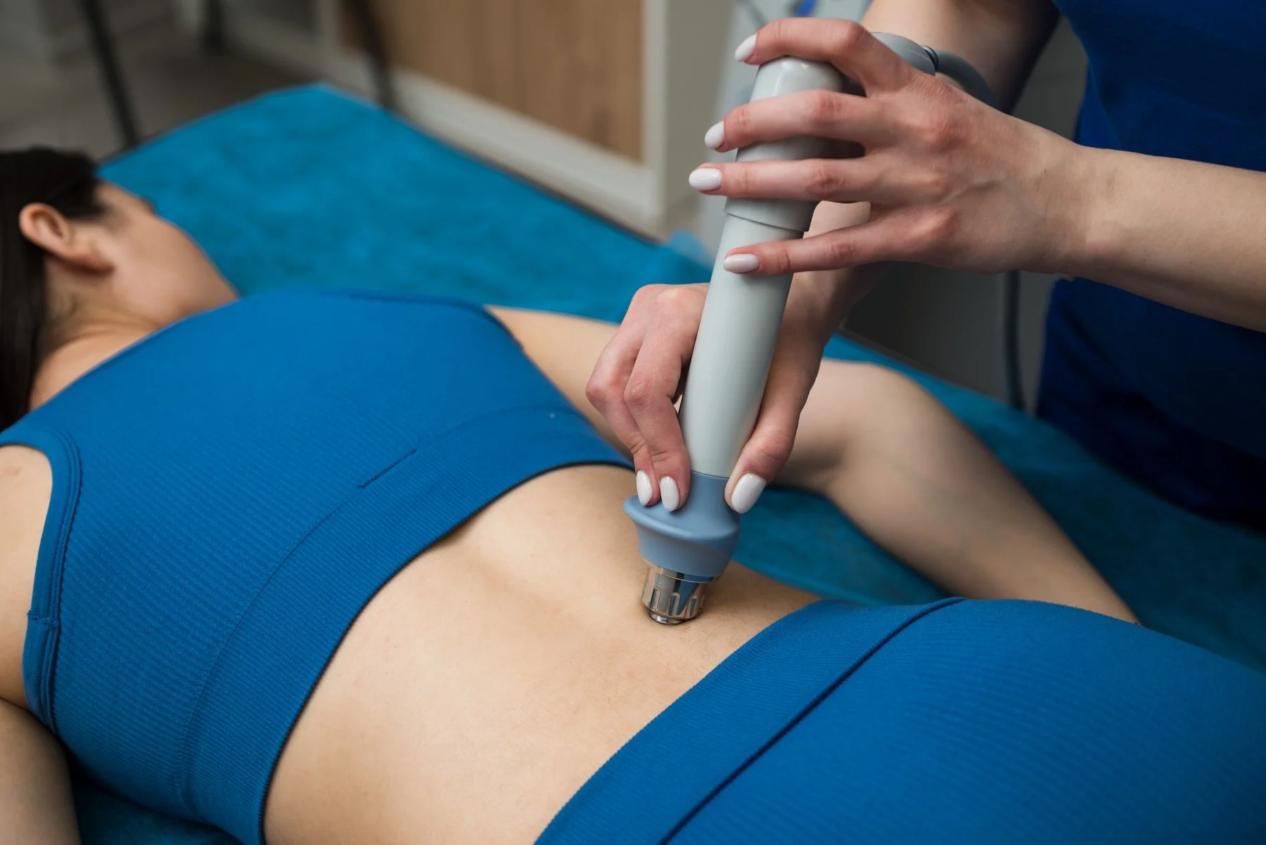 A person lying on a massage table receiving a treatment with a handheld device applied to their side, wearing a blue sports outfit.