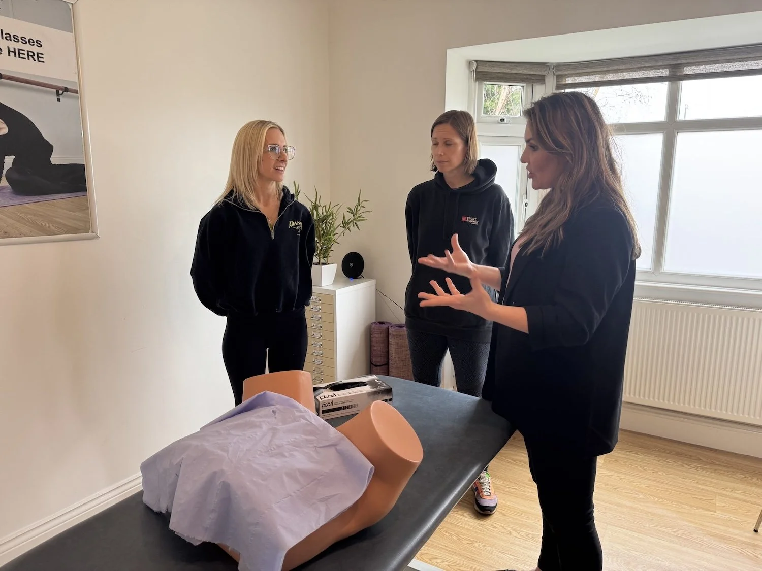 Three women standing around a training pelvis mannequin on a table, engaging in a discussion.