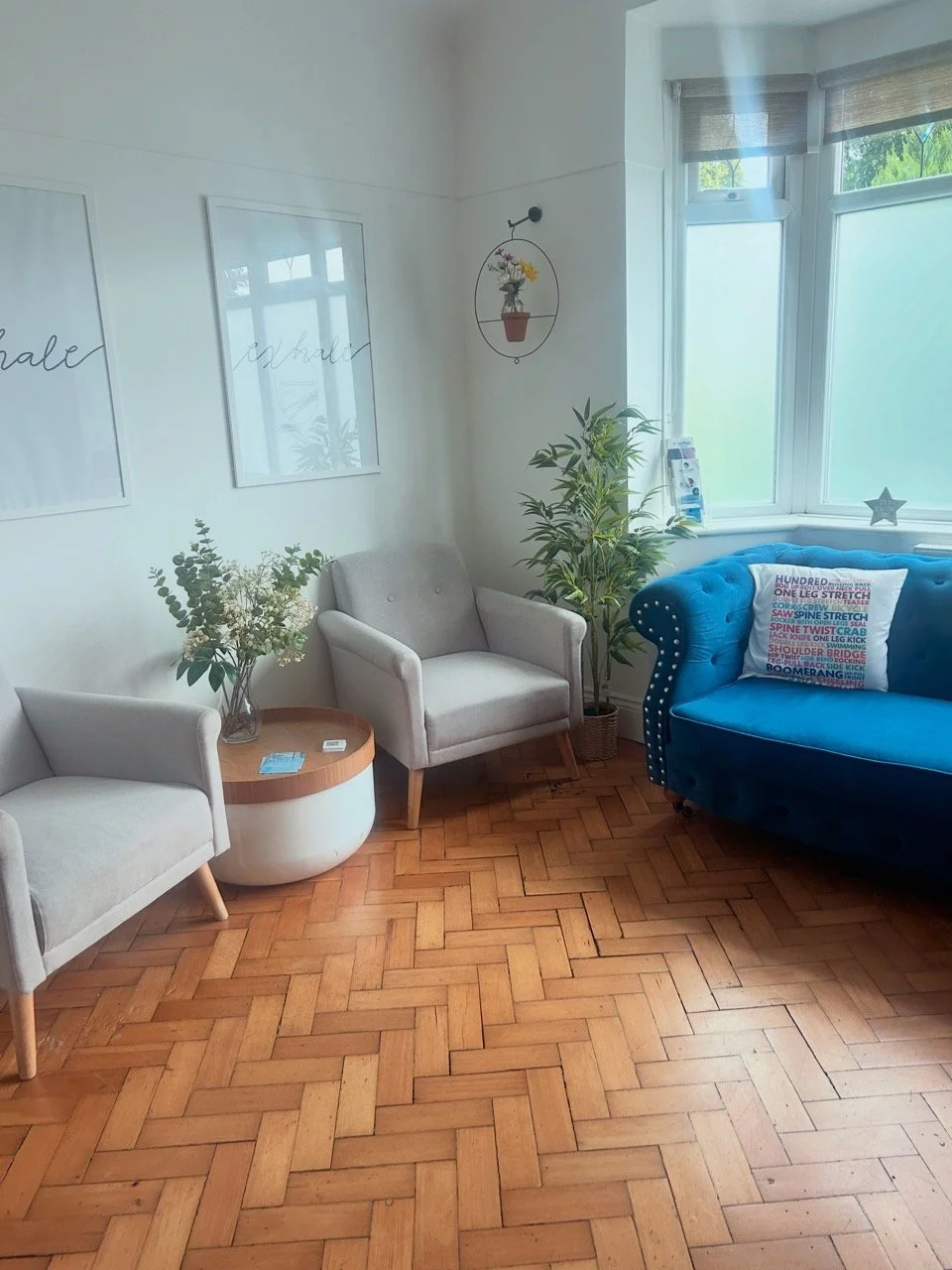 A cosy waiting room corner with two light grey armchairs, a small round side table, a blue velvet sofa with a colourful pillow, potted plants, framed wall art, a window with blinds, and hardwood flooring.