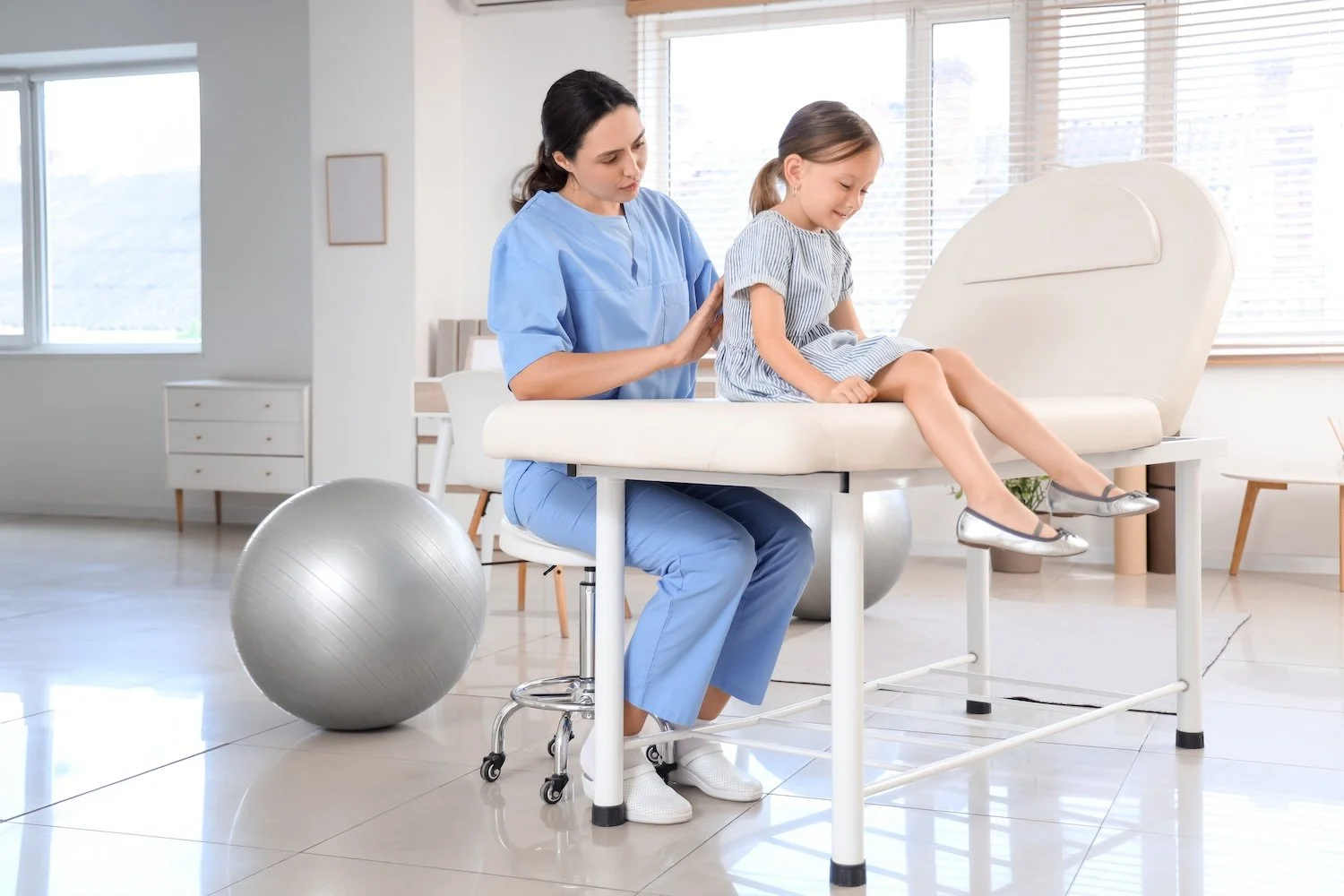 A female physiotherapist wearing blue scrubs, assists a young girl sitting on an examination table in a bright, modern clinic. The girl appears comfortable and is smiling, while the medical professional gently supports her.