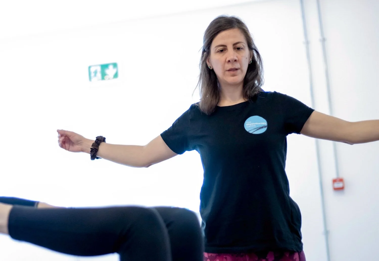 A female with shoulder-length brown hair wearing a black T-shirt with a blue logo for The Physiotherapy Centre, standing in a bright room with her arms outstretched, guiding a person performing a stretch.