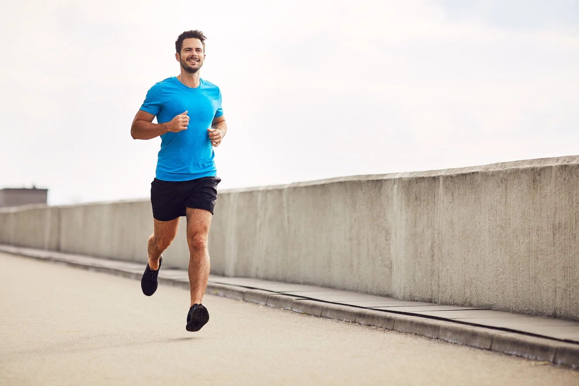 A man running outdoors on a paved path beside a concrete barrier wall, wearing a blue t-shirt, black shorts, and running shoes, smiling while jogging.