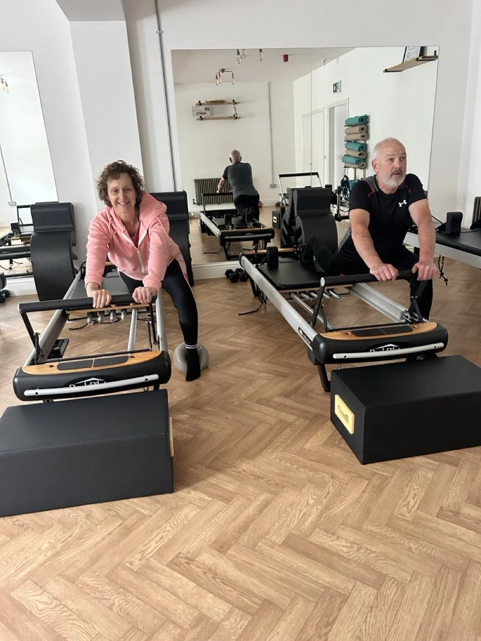 A male and a female take part in a reformer pilates session in a bright, light pilates studio.