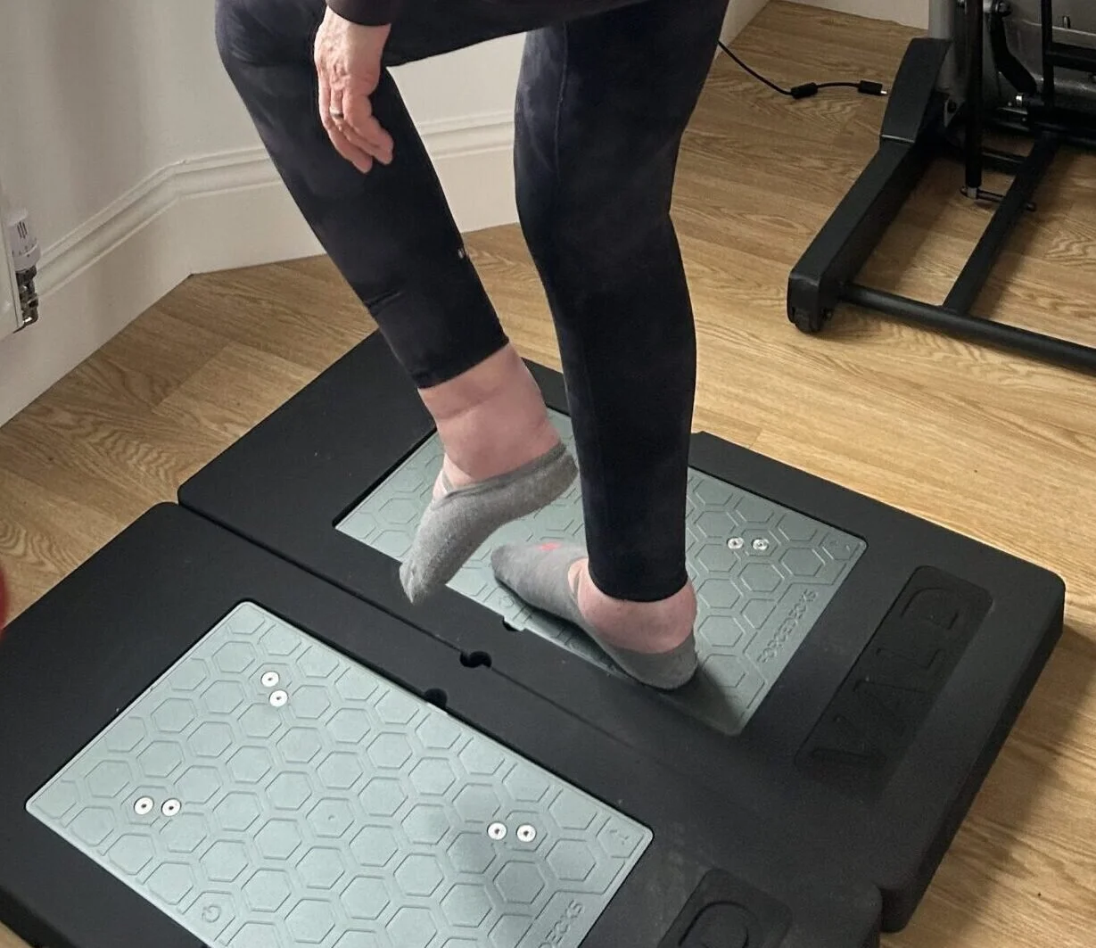 Person standing on a force plate in socks, with their left foot raised, in a room with wooden flooring.