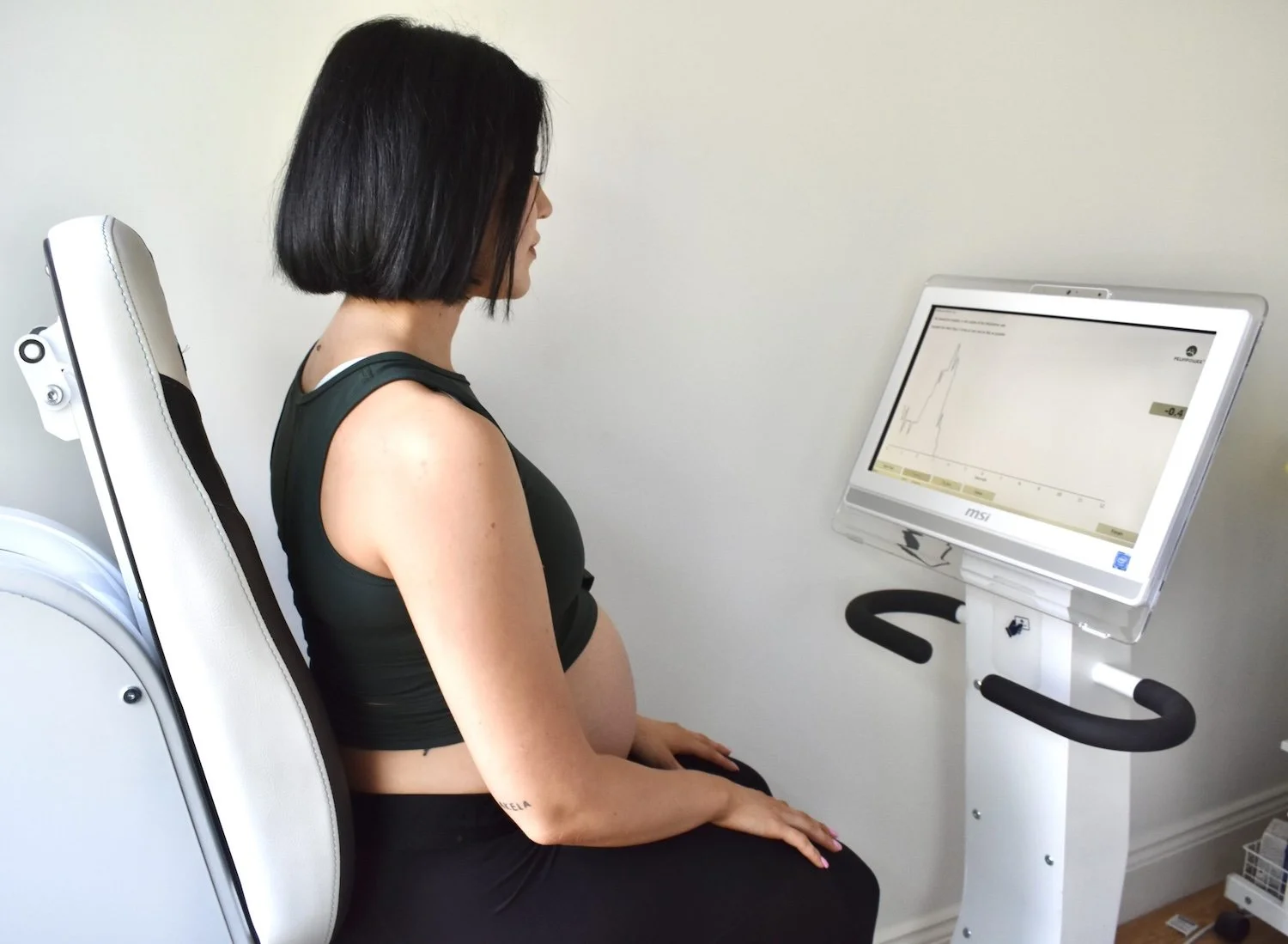A pregnant woman with short black hair and wearing a black sleeveless top is sitting on a medical chair while looking at a screen displaying a graph with post partum information.