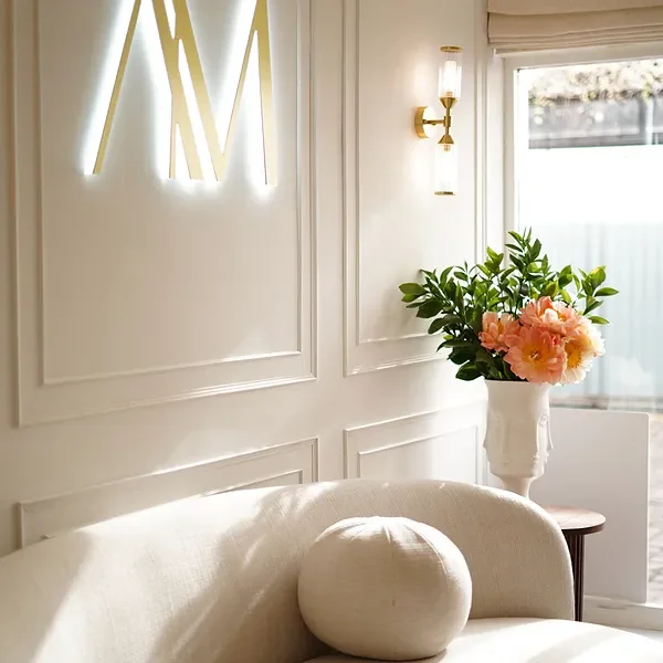 A cozy corner of a room with white walls, a beige sofa with a round pillow, a side table with a large white vase holding pink peonies and green foliage, a wall-mounted light fixture, and a window with sheer curtains.
