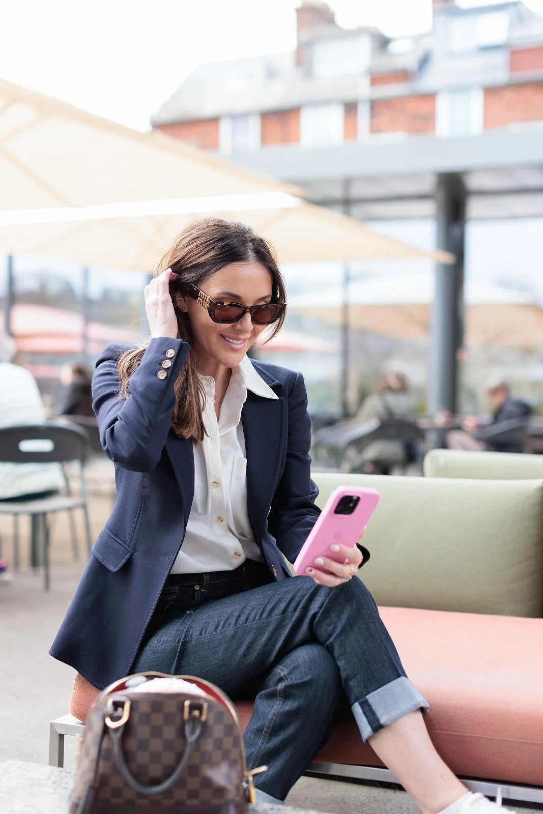 A woman sitting outdoors at a cafe, wearing sunglasses, a navy blazer, white blouse, and jeans, looking at her pink smartphone with a smile.
