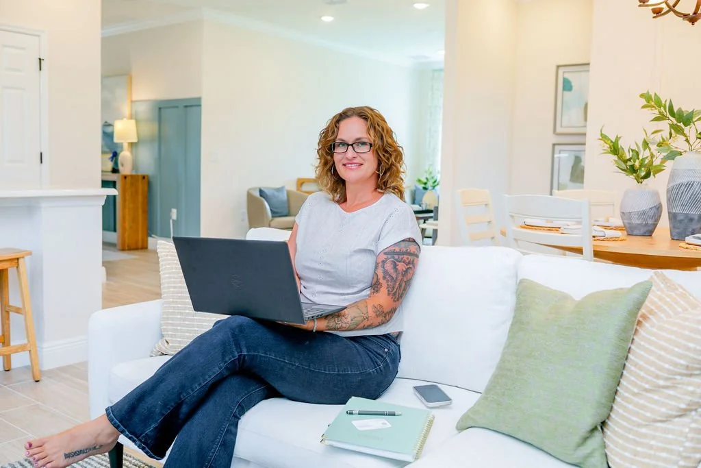 Woman with curly hair and glasses sitting on white sofa with laptop, in a bright, modern living room decorated with plants and artwork.