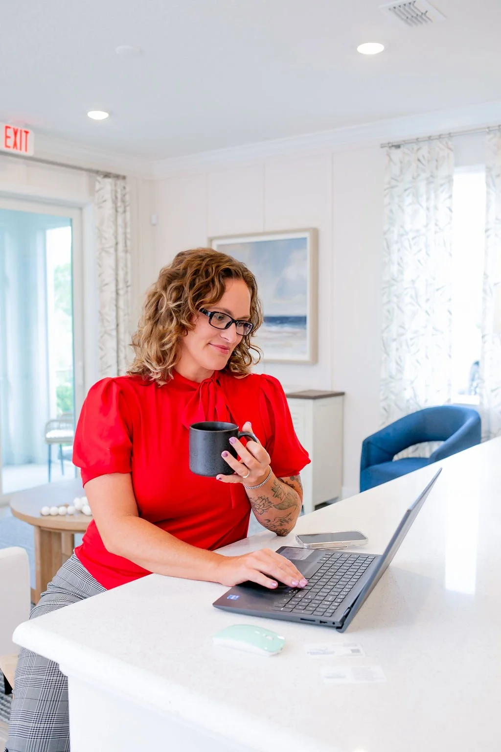 A woman with curly hair, glasses, and tattoos on her arm, wearing a red blouse, holding a black mug, working on a laptop at a kitchen island.