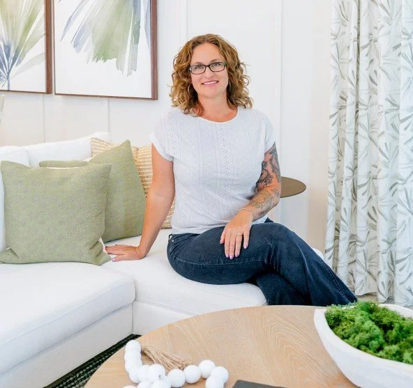A woman with glasses and curly hair sitting on a white couch with green and beige pillows, smiling in a bright living room with modern art and patterned curtains.