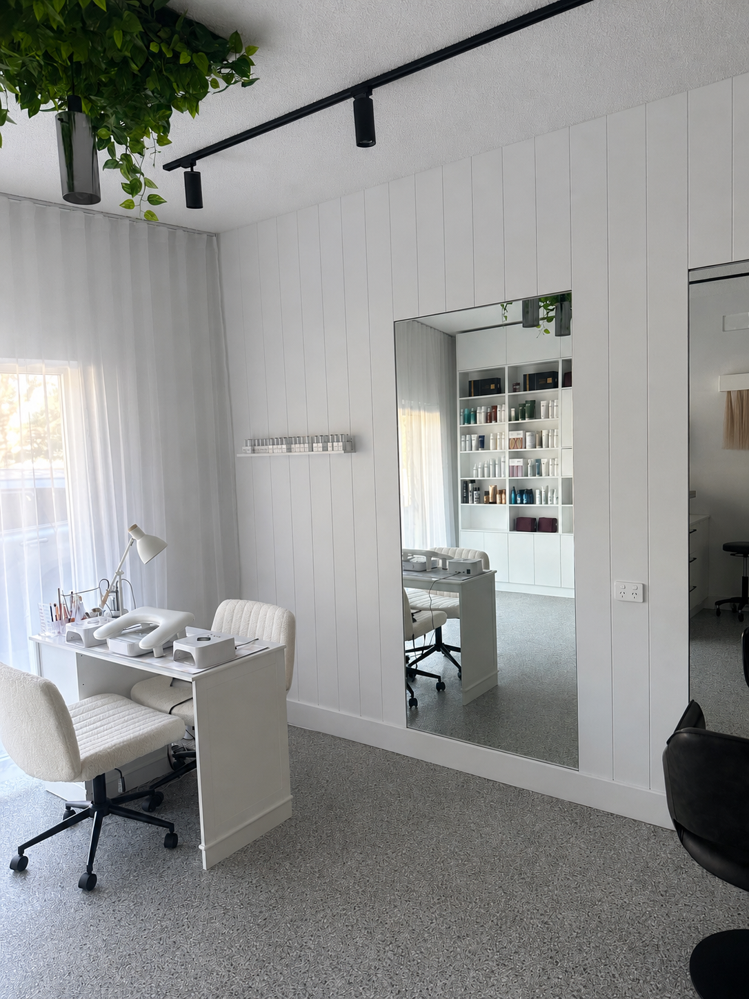 Interior of a modern beauty or nail salon with white furniture, a desk with beauty tools and equipment, a large mirror, shelves with products, and a gray carpeted floor.