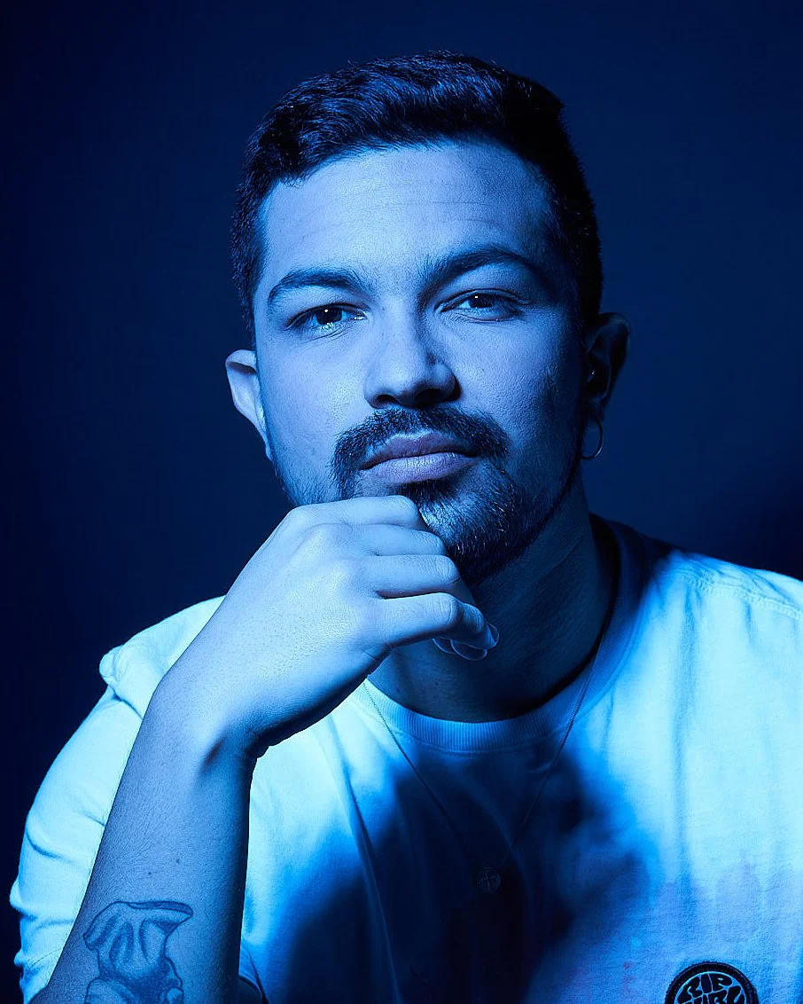 Retrato de um homem com barba, cabelo curto, em um fundo escuro, com iluminação azul. Ele está com a mão no queixo e olhando diretamente para a câmera.