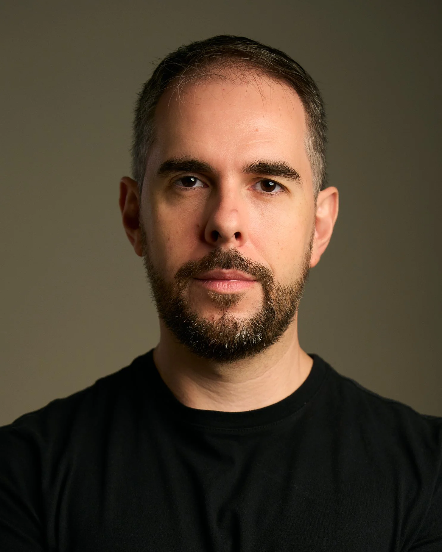 Retrato de um homem com barba e cabelo castanho claro, usando camiseta preta, em fundo neutro.
