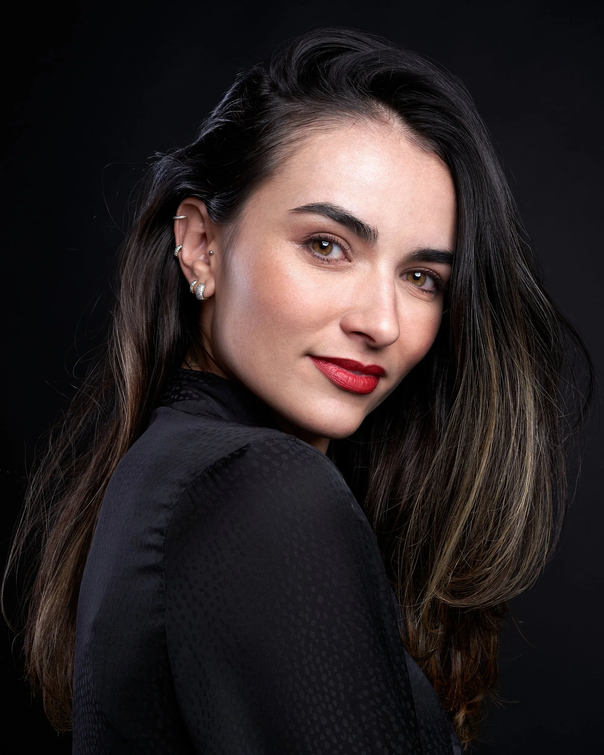 Retrato de uma mulher com cabelo castanho escuro e olhos castanhos, usando batom vermelho, brincos de argola e uma blusa preta, contra fundo preto.