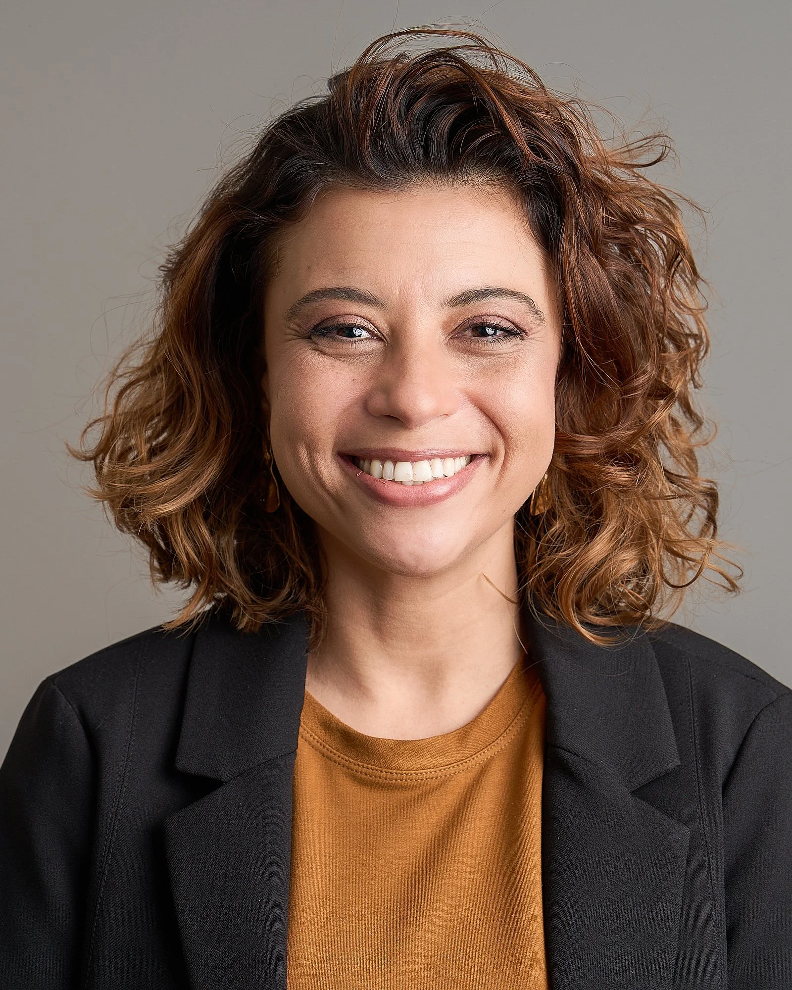 Retrato de uma mulher sorridente com cabelo cacheado marrom, usando blazer preto e blusa amarela, contra fundo cinza claro.