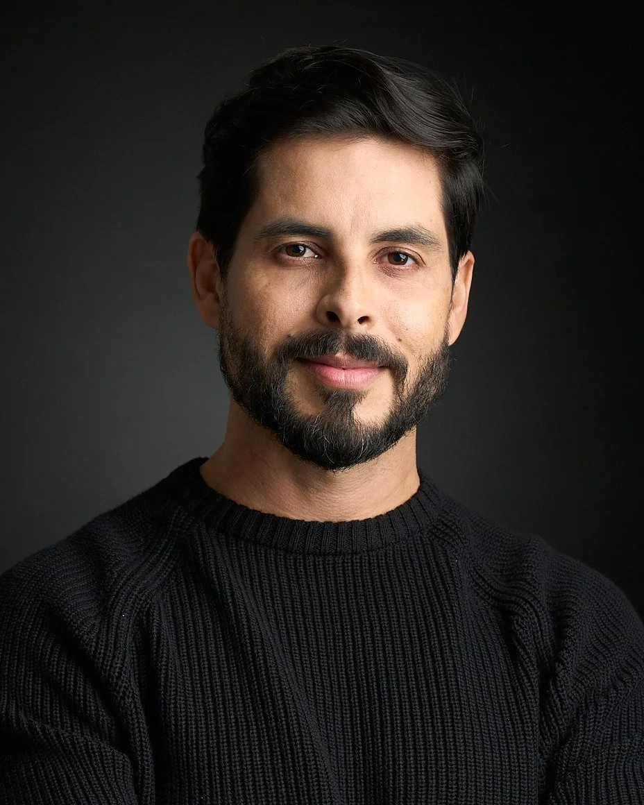 Homem com barba, cabelos escuros e cabelo penteado, usando suéter preto, posando contra fundo escuro.