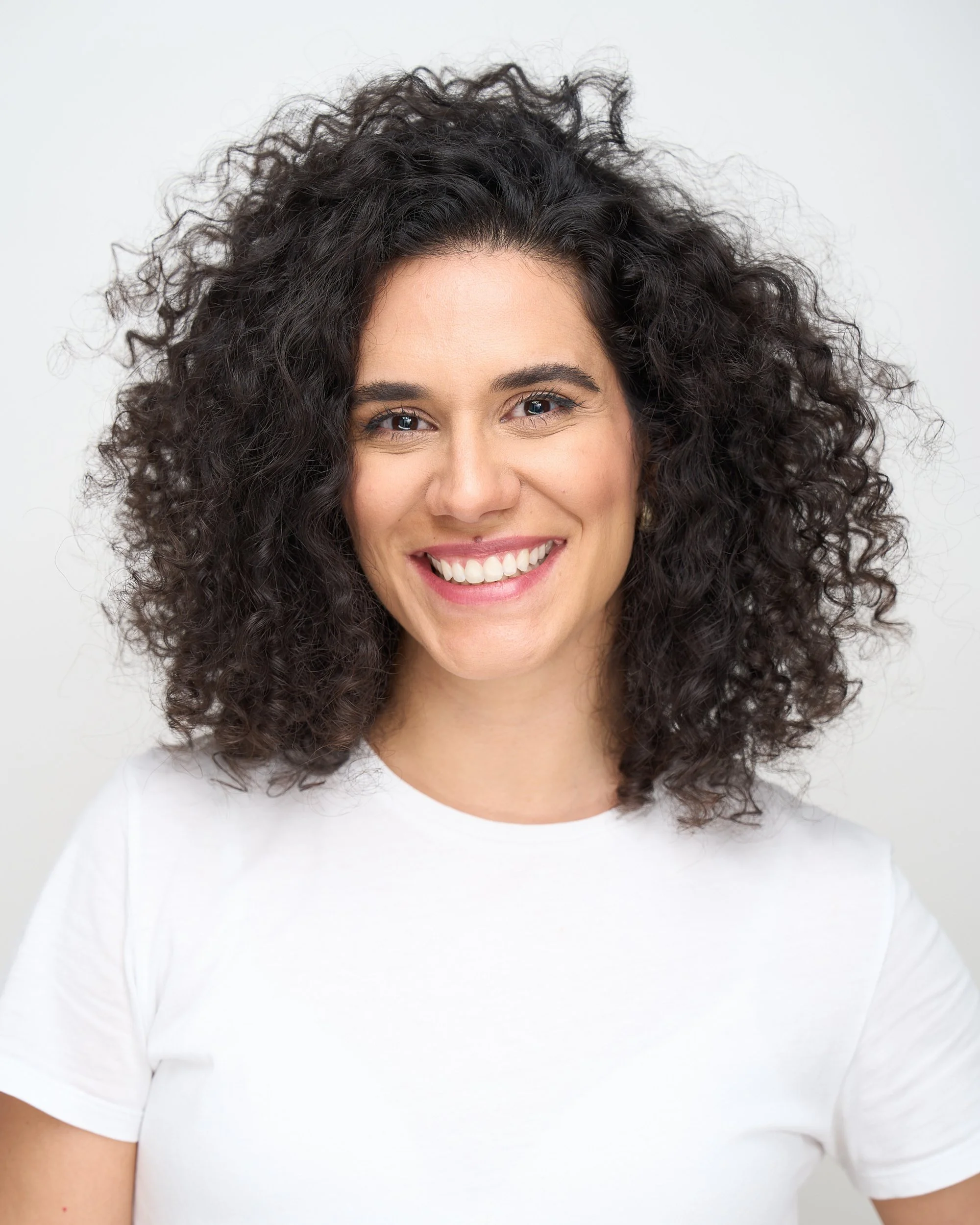 Mulher sorrindo, cabelo cacheado escuro, vestindo camiseta branca, fundo branco.