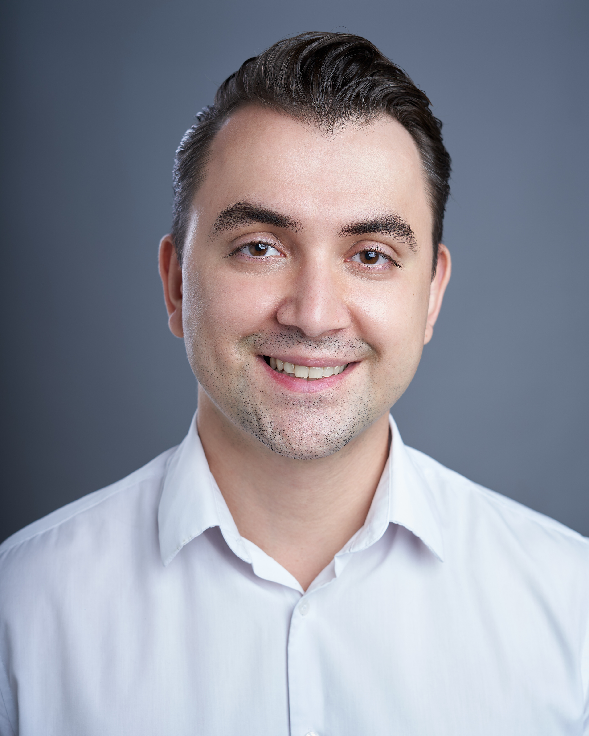 Retrato de um homem sorridente usando camisa branca, com cabelo escuro e penteado, contra fundo cinza.