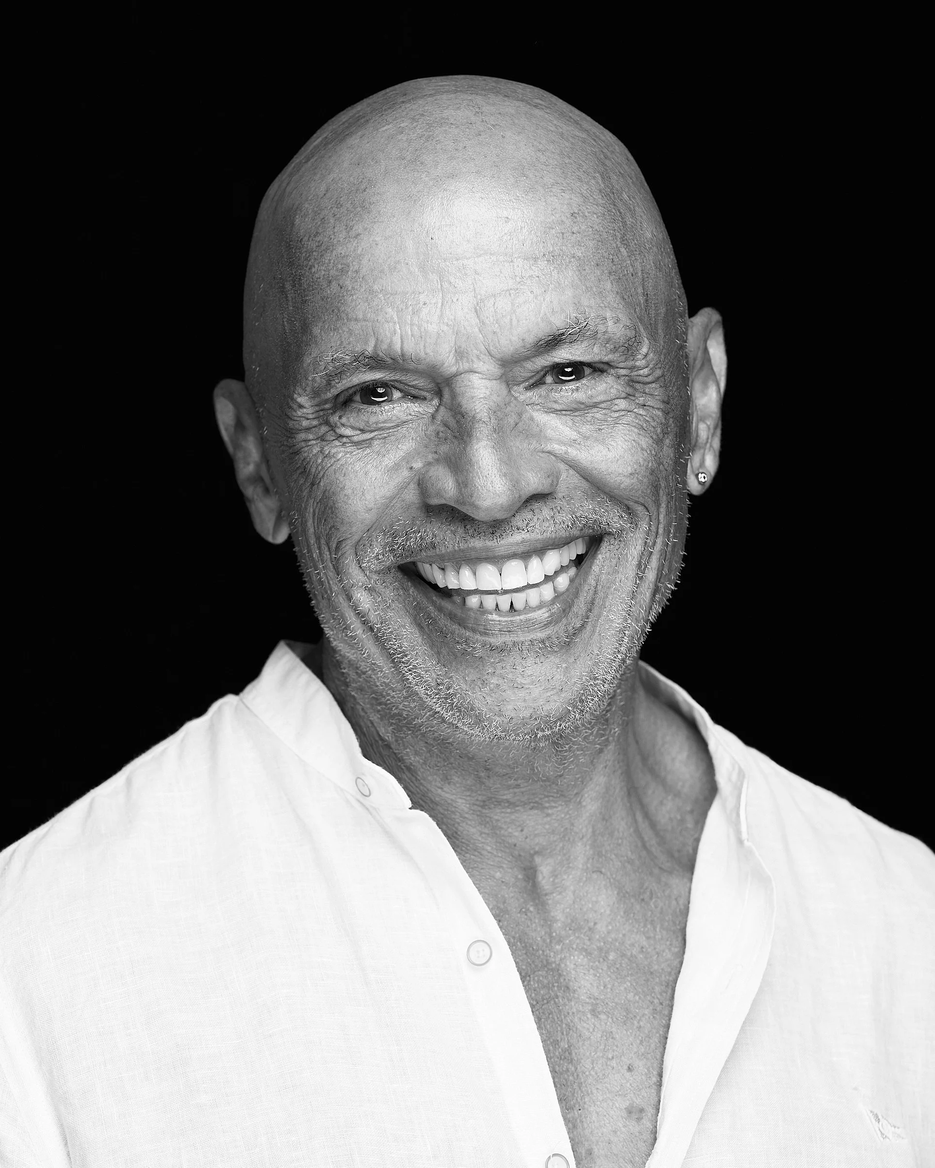 Retrato preto e branco de um homem idoso sorridente, com cabeça careca, barba grisalha, usando camisa branca.