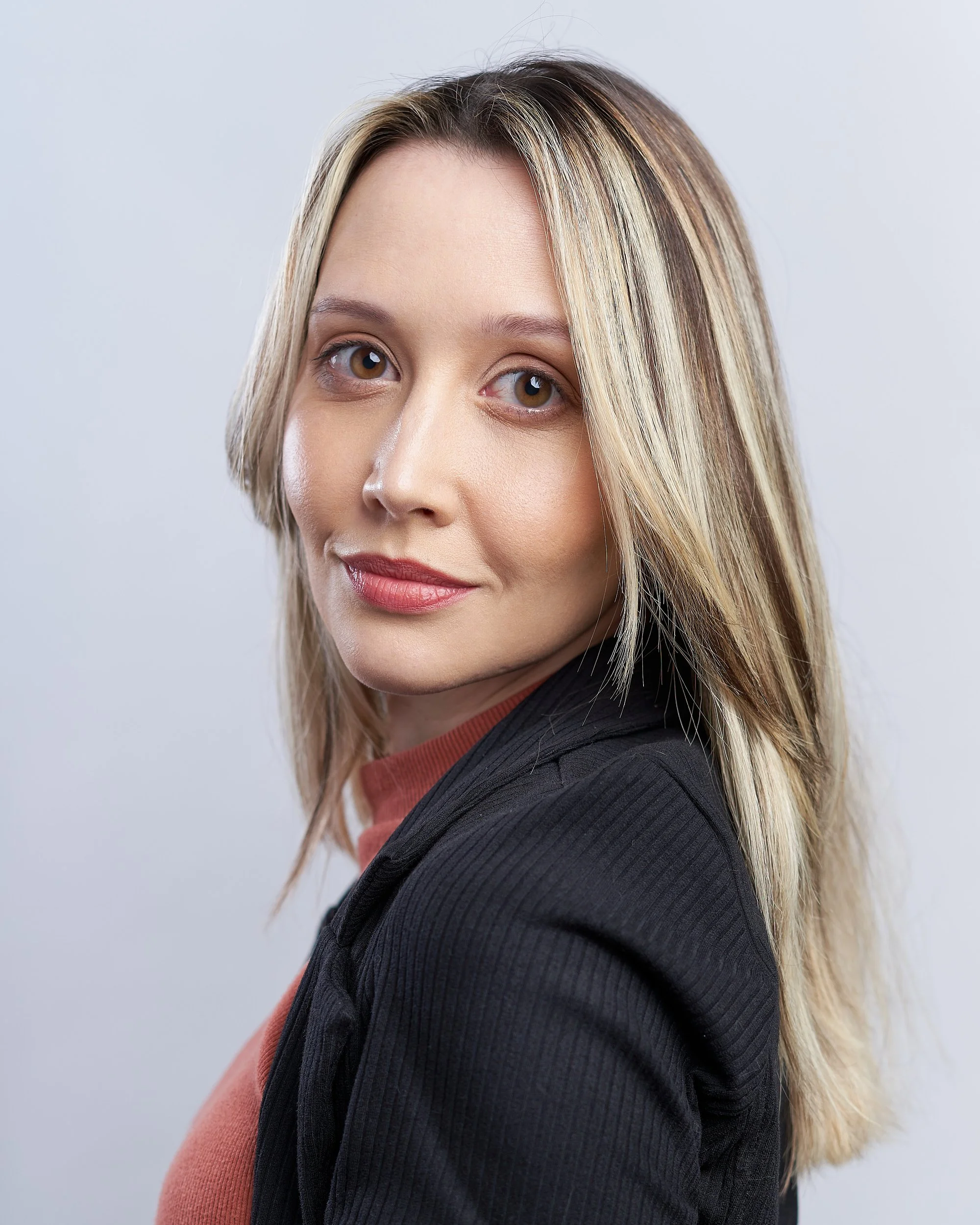 Retrato de mulher com cabelo castanho claro e mechas loiras, usando blazer preto e suéter marrom, sorrindo levemente, com fundo cinza claro.
