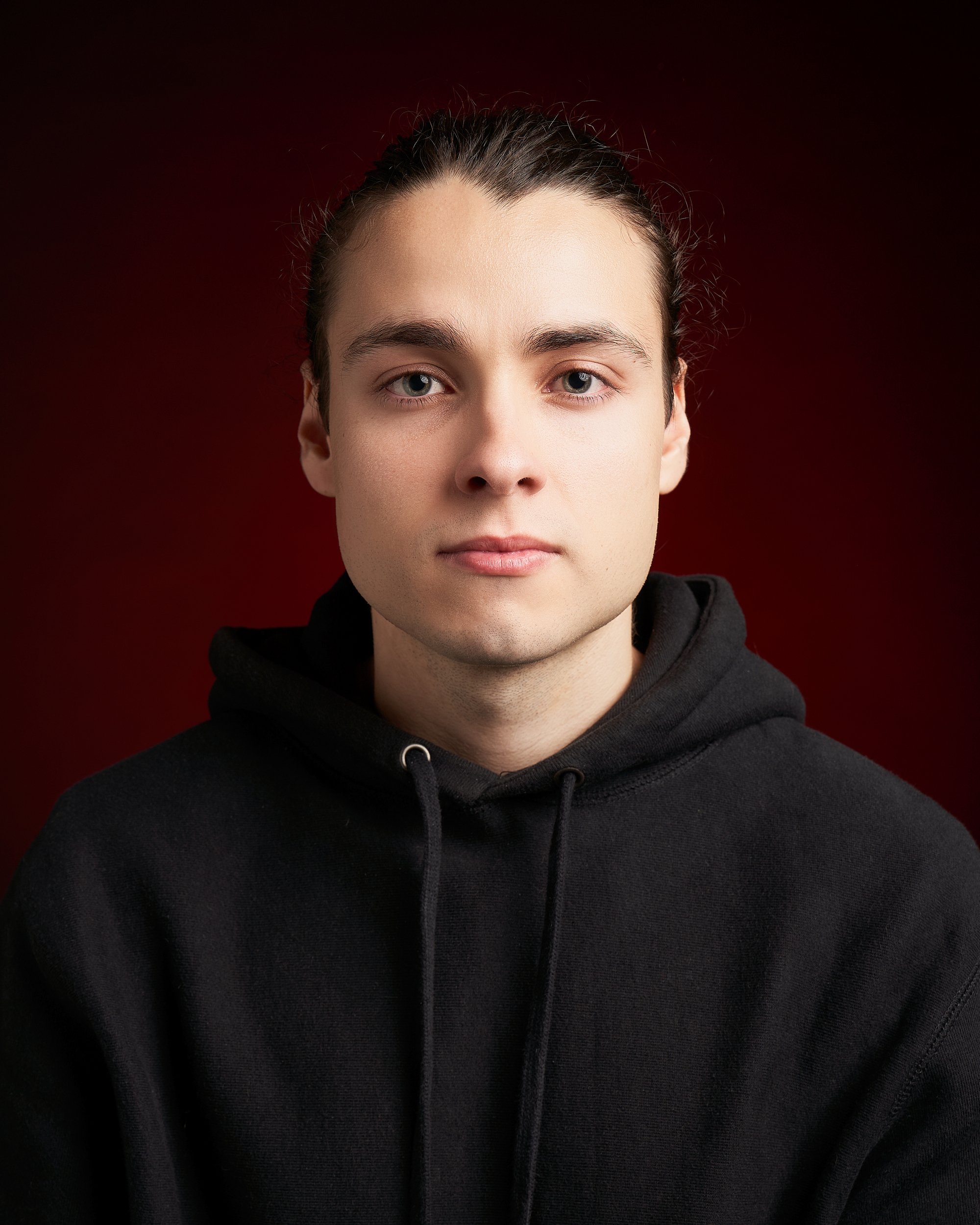 Retrato de um jovem com cabelo penteado para trás, expressão neutra, usando blusa preta, fundo vermelho escuro.