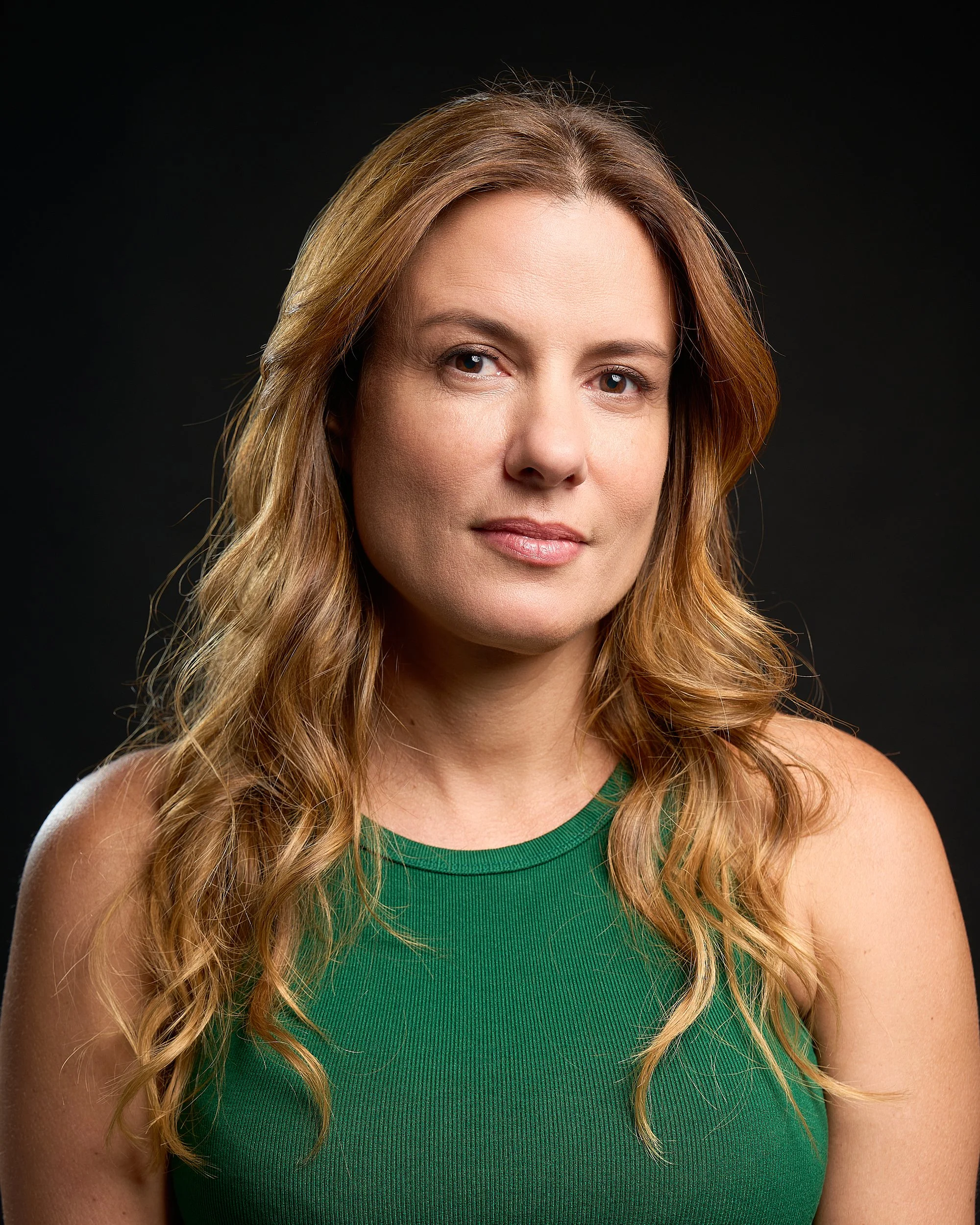 Retrato de uma mulher de cabelos longos e castanhos, usando uma blusa verde, com fundo preto.