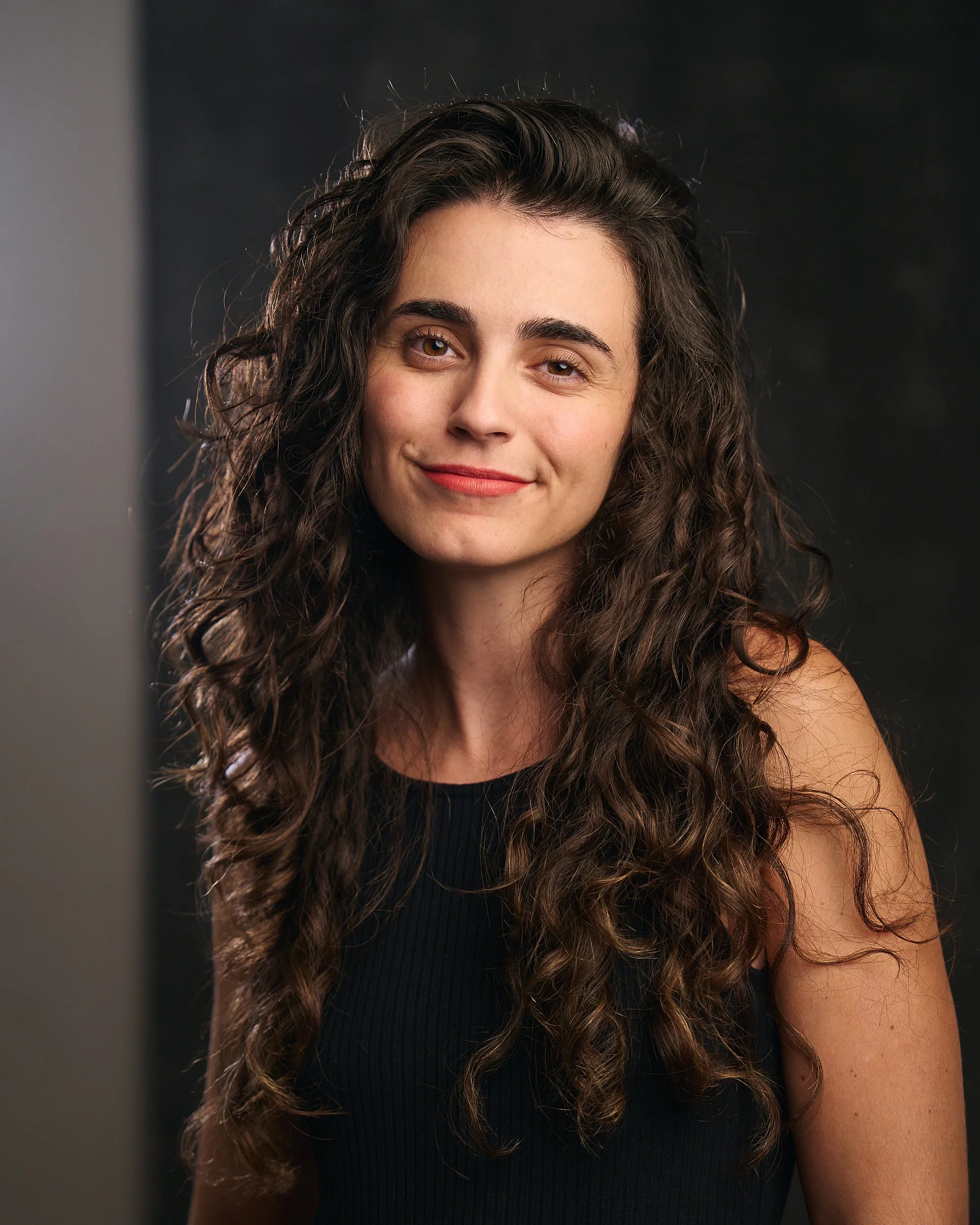 Mulher sorridente com cabelo cacheado longo e escuro, usando roupa preta, em ambiente com fundo escuro.