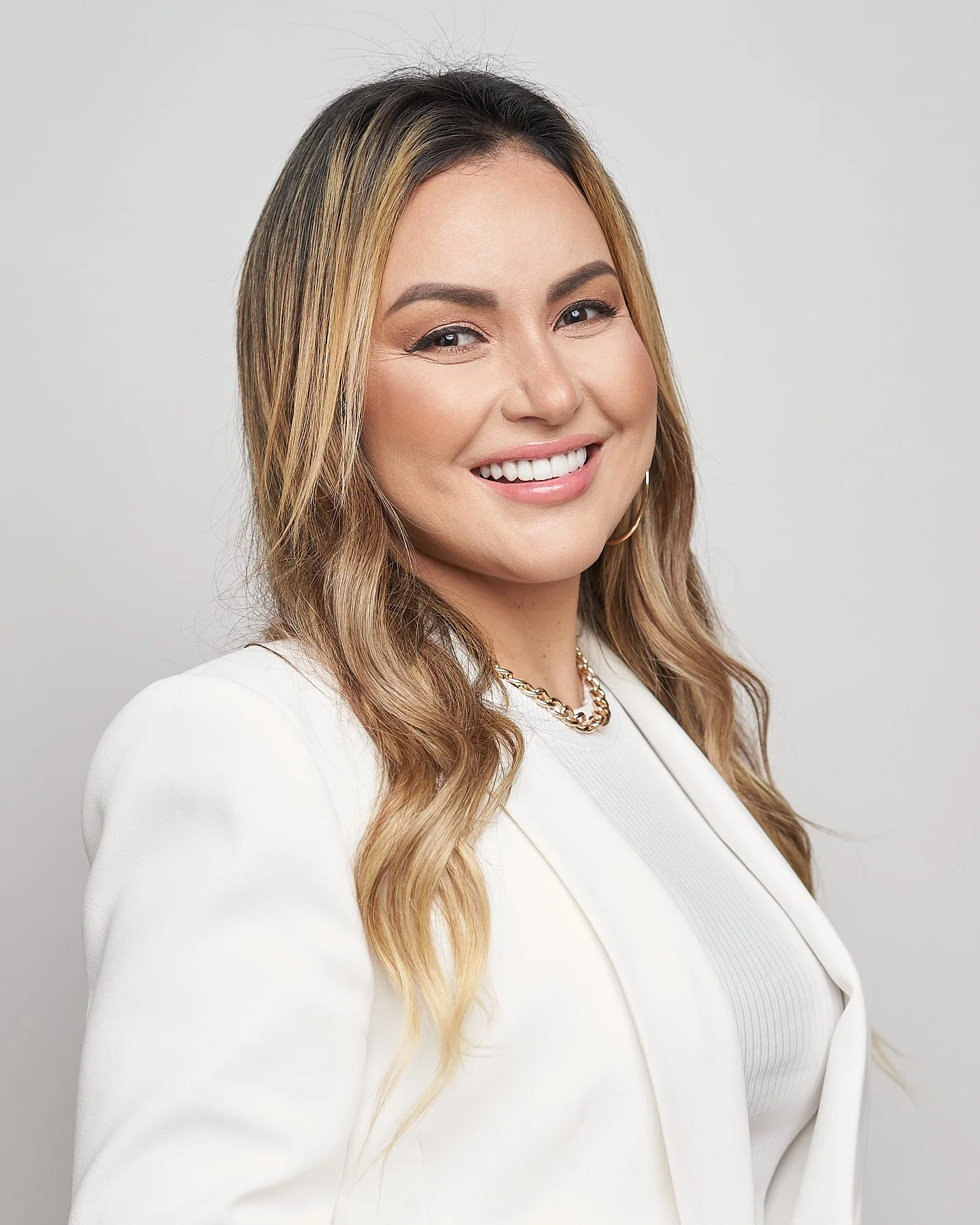 Mulher sorrindo com cabelo castanho claro, usando brinco de argola e colar de corrente dourada, vestindo blazer branco.