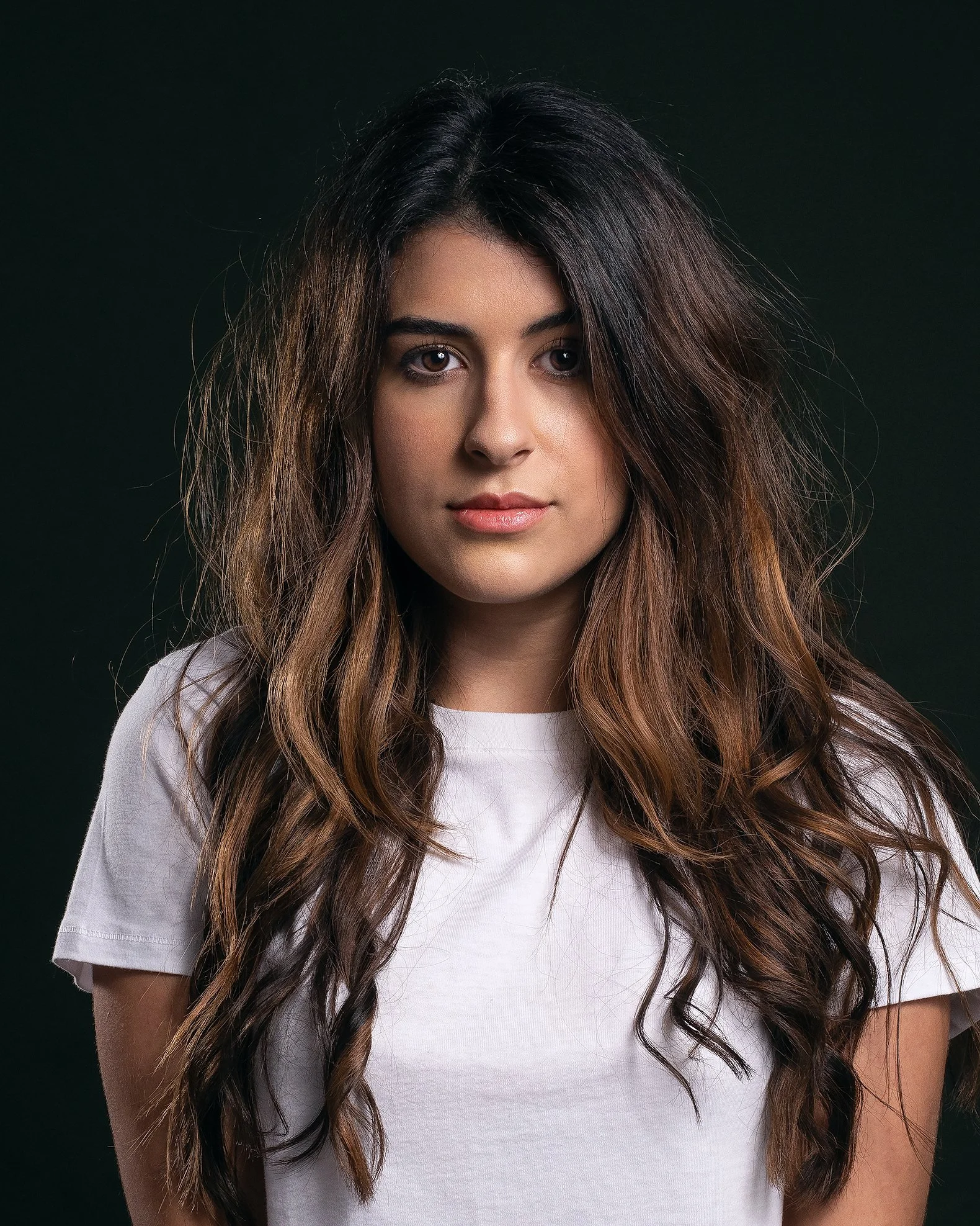 Retrato de uma jovem mulher com cabelo castanho, ondulado, vestindo uma camiseta branca, fundo escuro