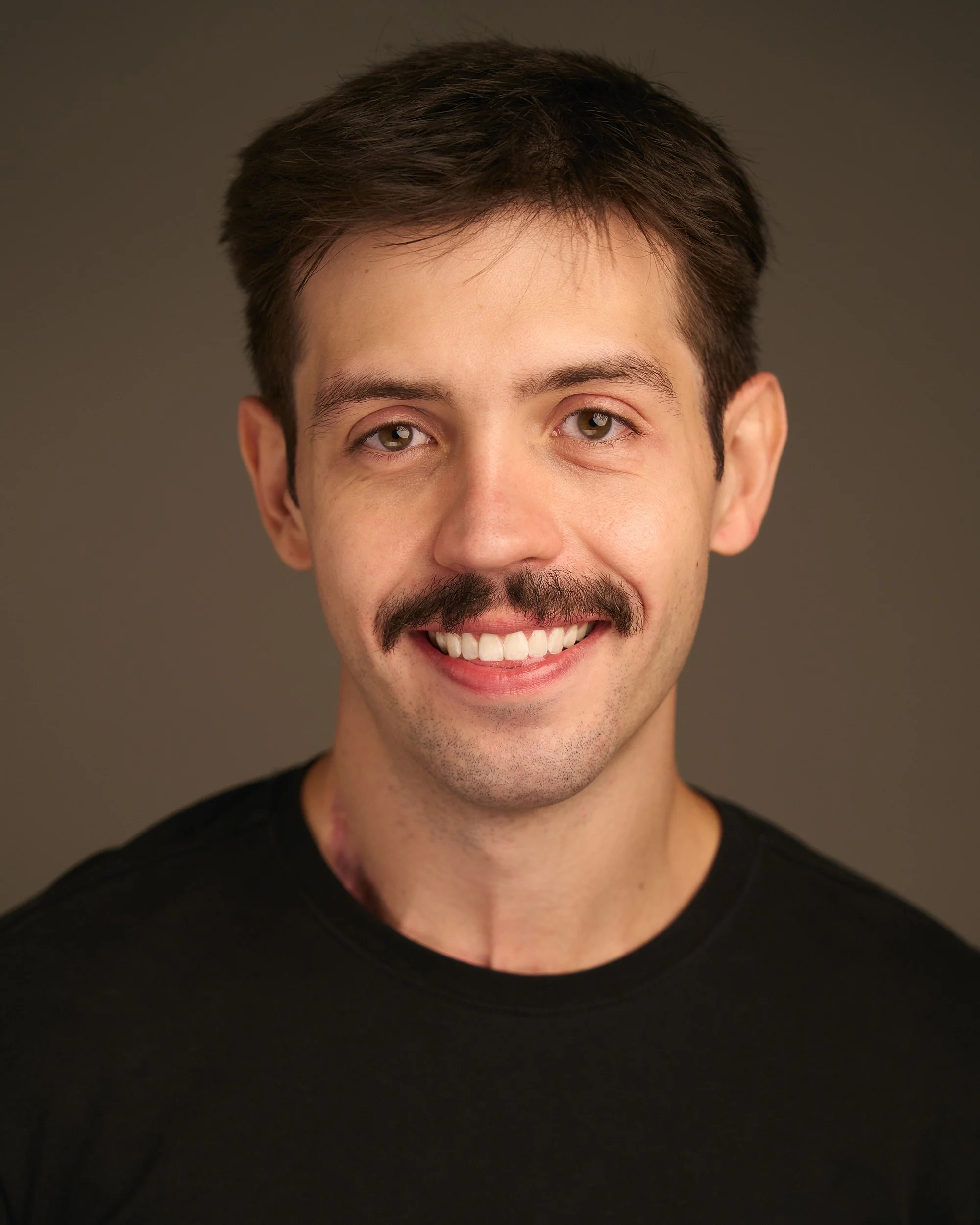 Retrato de um homem sorridente de cabelo castanho escuro e barba leve, usando uma camiseta preta, contra fundo cinza