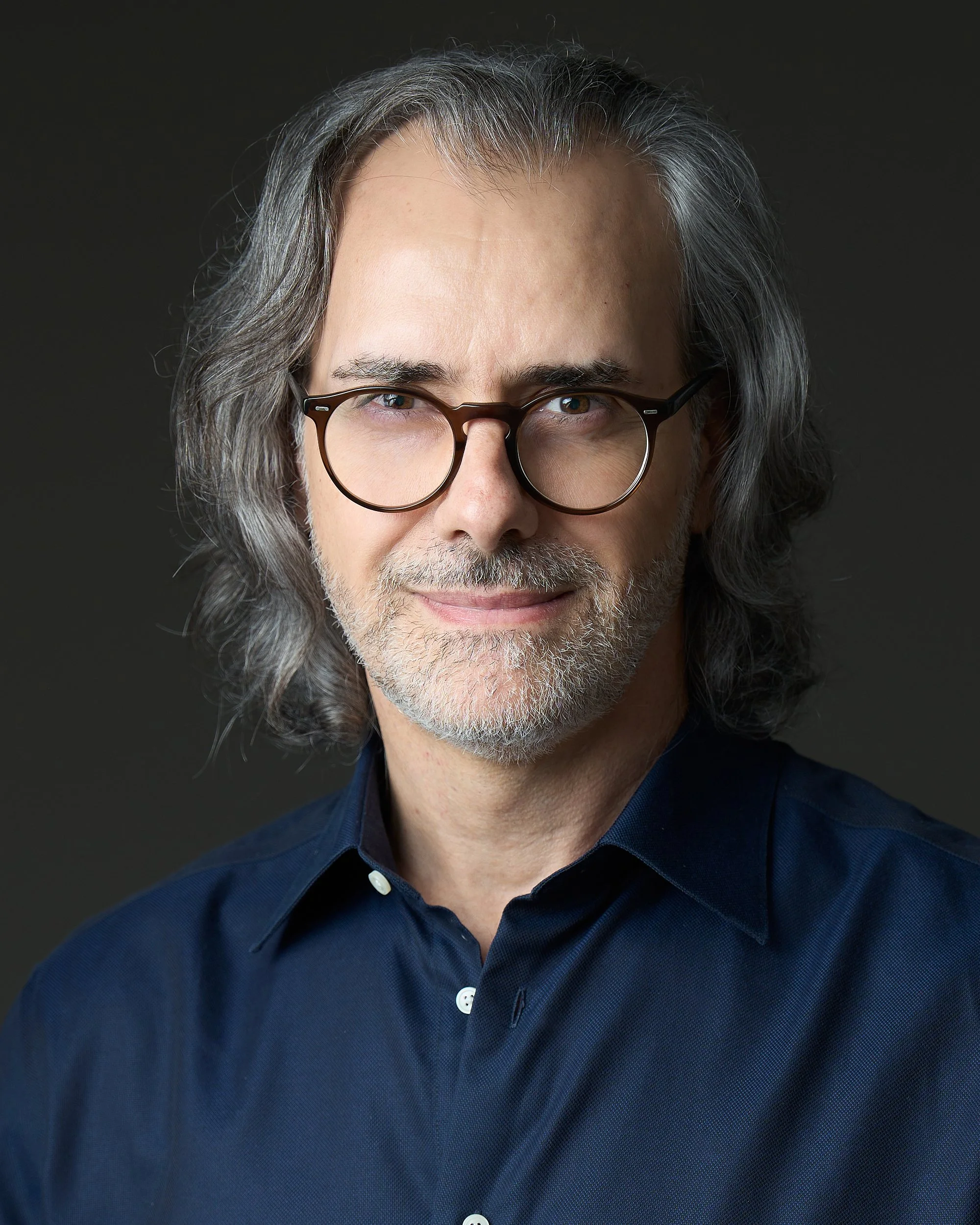 Retrato close-up de um homem de meia-idade com cabelo grisalho, barba por fazer, óculos de armação grossa, vestindo camisa azul escuro, com fundo preto.
