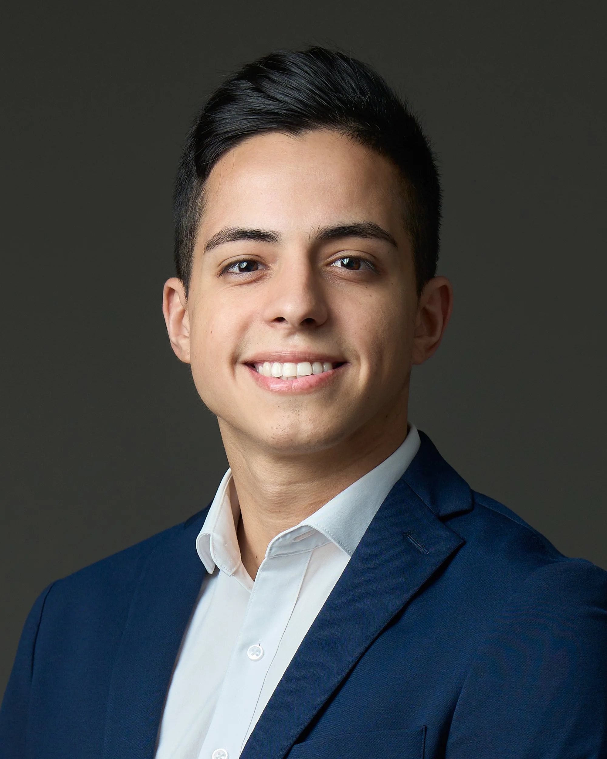 Retrato de um jovem homem sorridente com cabelo escuro e bem penteado, vestindo terno azul escuro e camisas branca, contra fundo cinza escuro.