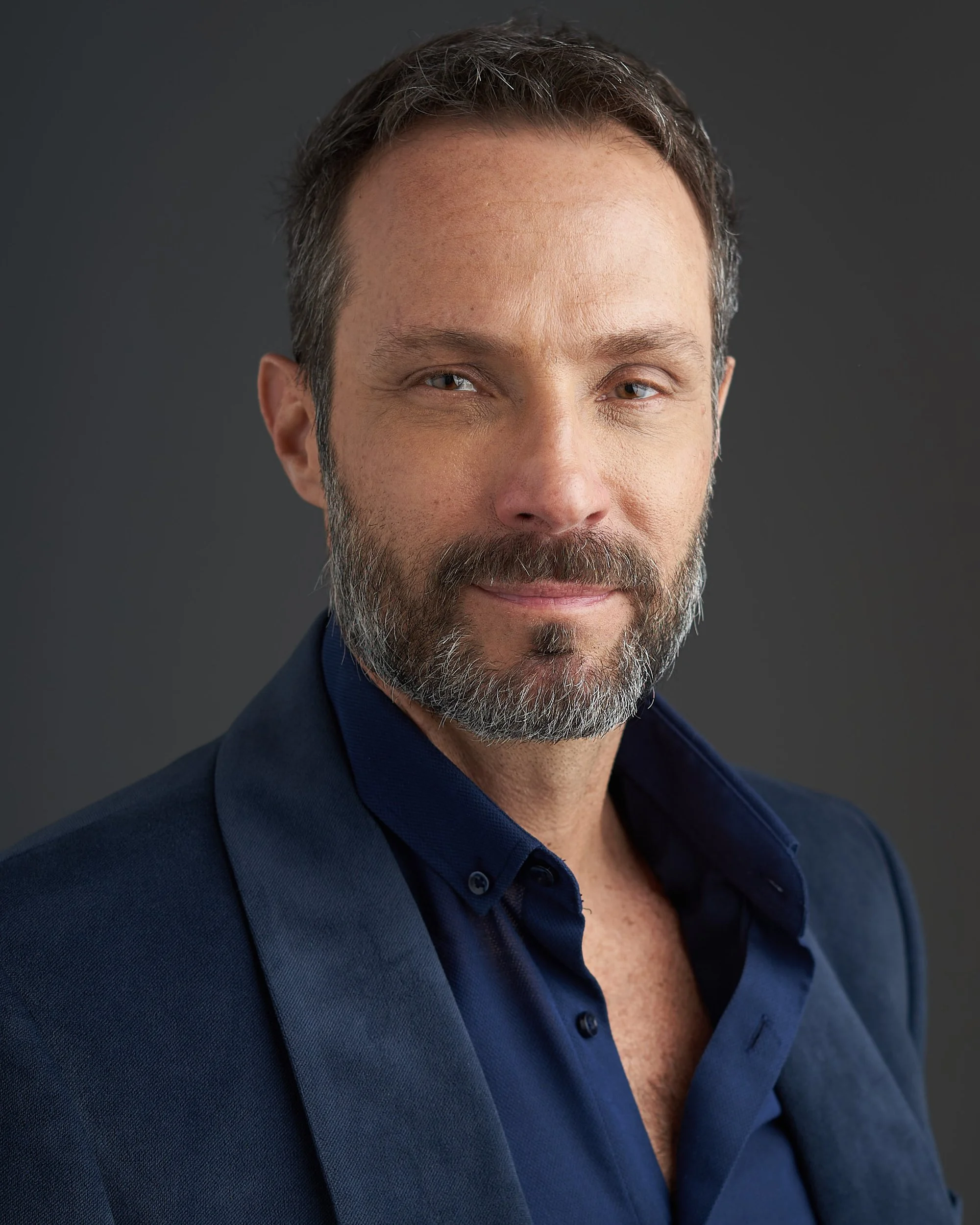 Retrato de um homem com barba e cabelo castanho, usando uma camisa azul escura, sorrindo levemente, contra um fundo cinza escuro.