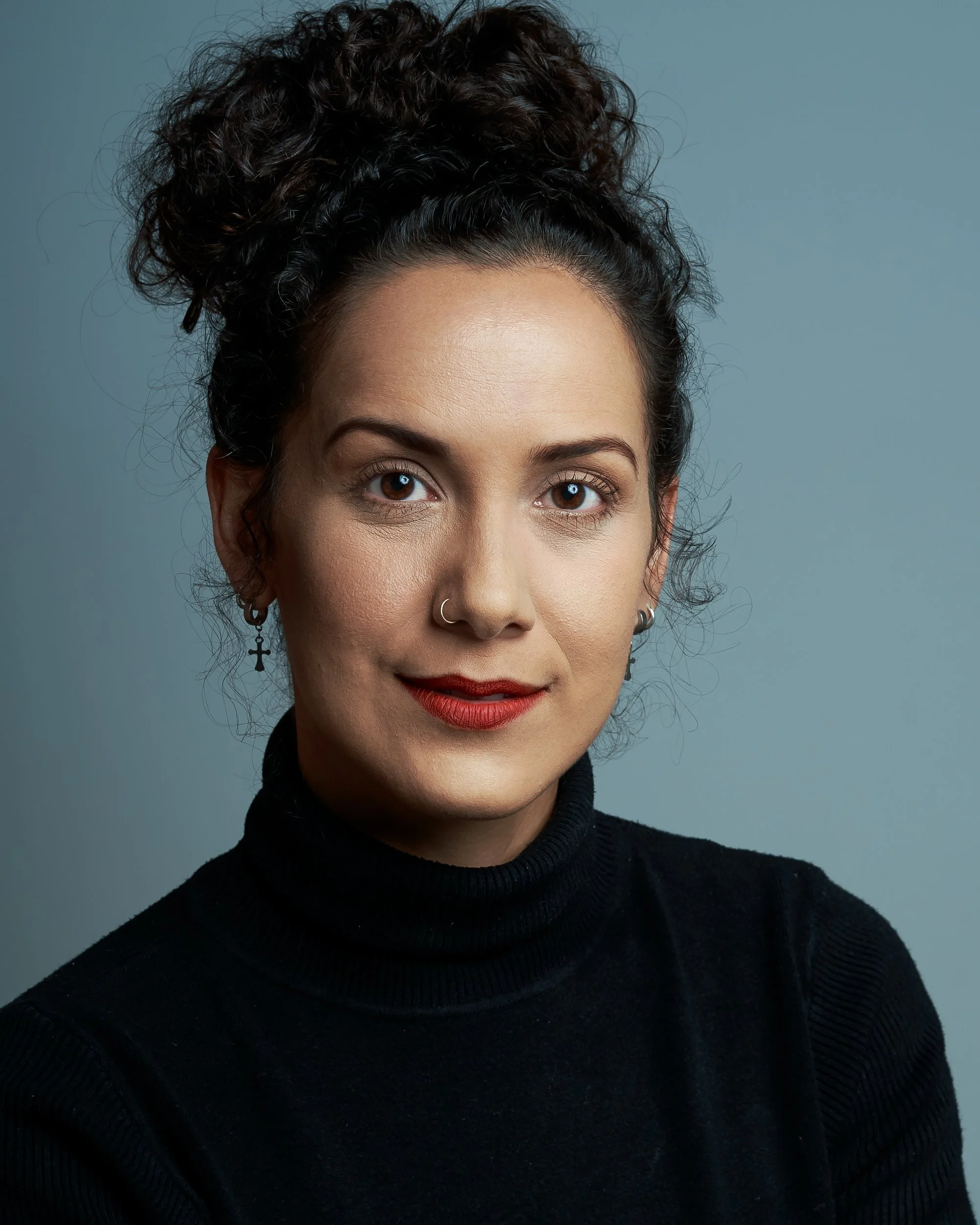 Retrato de uma mulher com cabelo cacheado preso em um coque alto, usando brincos com cruz e um sutiã de gola alta preto, contra um fundo cinza claro.