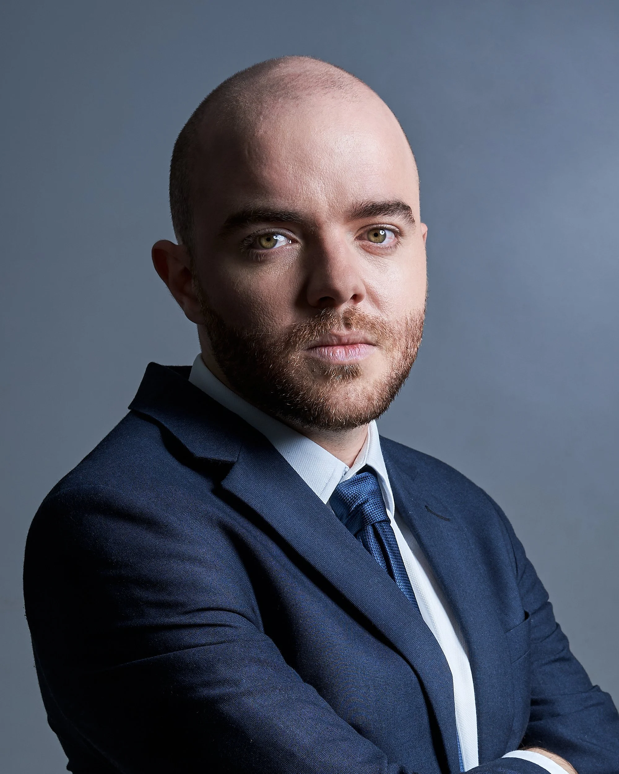 Homem de terno azul escuro e gravata azul, com cabelo raspado e barba, olhando para a câmera, contra fundo cinza.