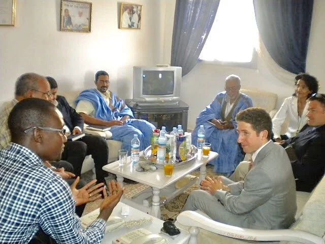 A diverse group of people, including men and women, sitting around a table drinking and talking in a living room.