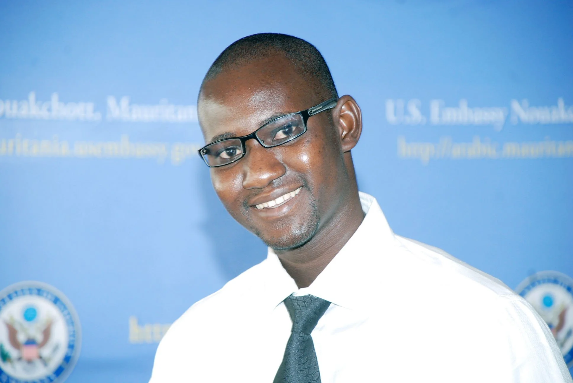 A man with dark skin, wearing glasses, a white shirt, a black tie, and a dark coat, smiling and standing in front of a blue backdrop with logo and text related to U.S. Embassies and the United States.
