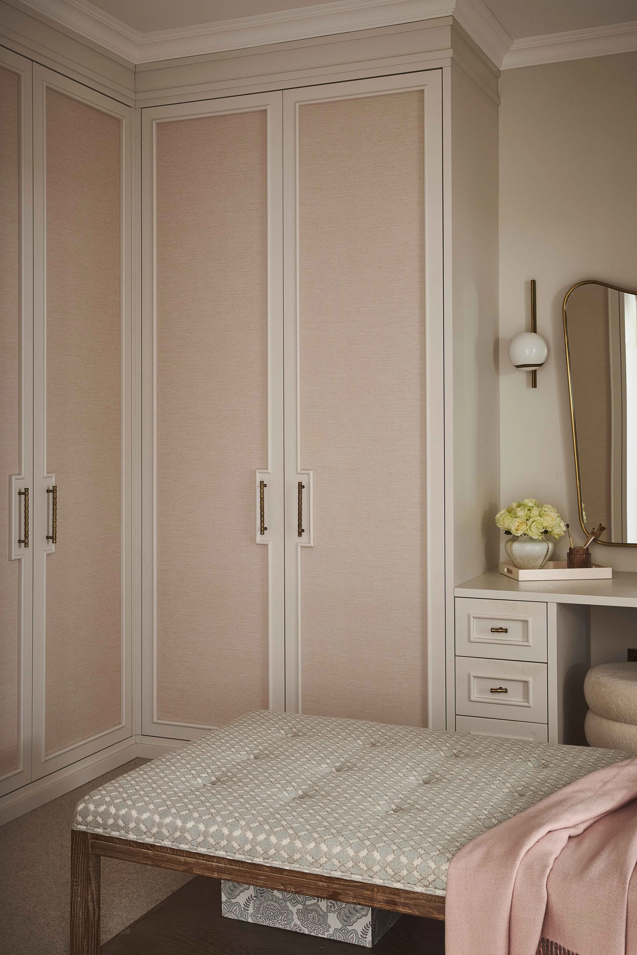 A bedroom corner with a white wardrobe, a white nightstand with drawers, a lamp, a mirror, a bouquet of white flowers, and a bed with a textured mattress and pink blanket.