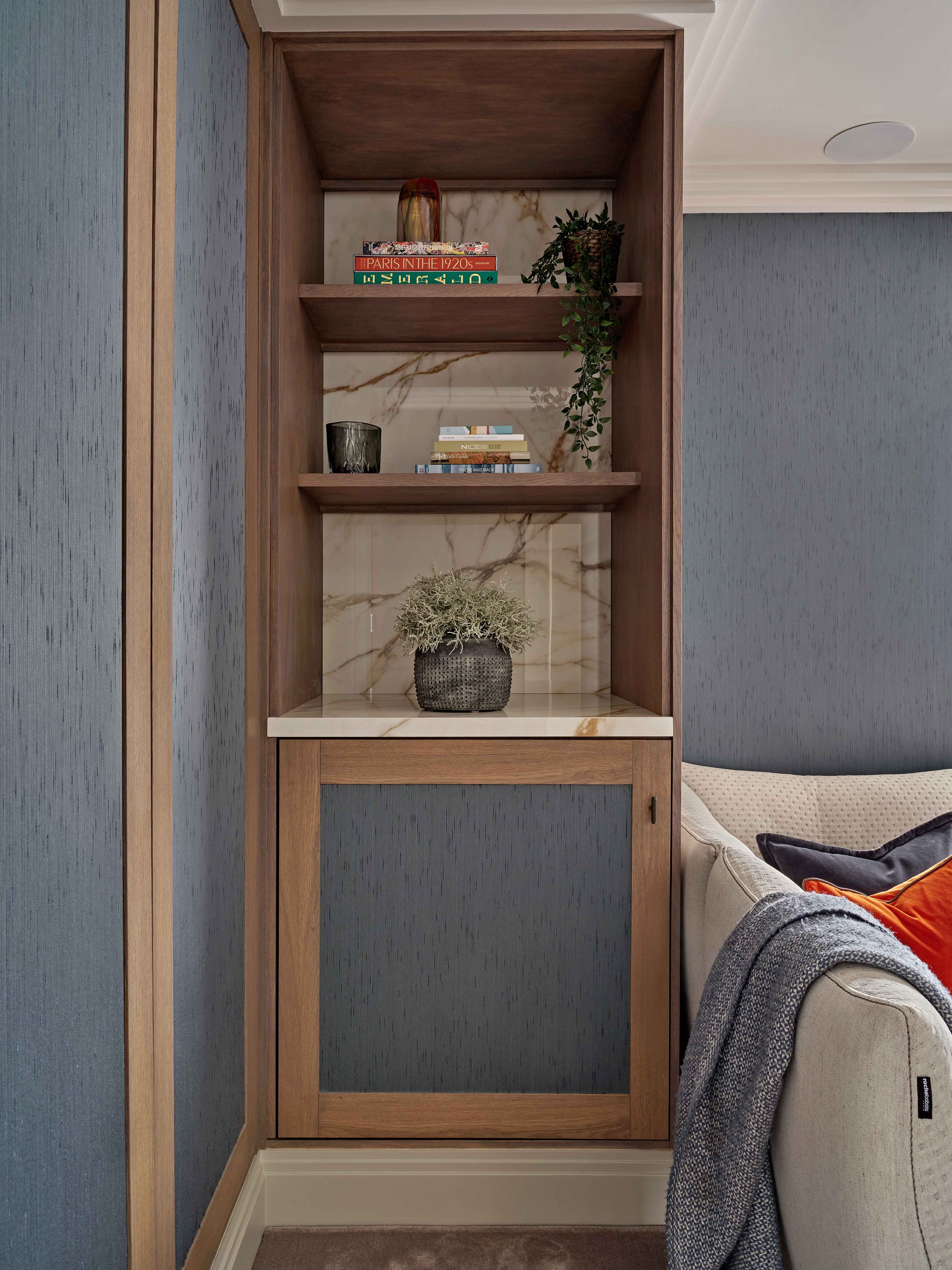 A wooden shelf unit built into a wall, with three shelves. The top shelf has a decorative vase and three books, one titled 'Paris in the 1920s'. The middle shelf has a small potted plant, a black cup, and some books. The bottom shelf has a decorative