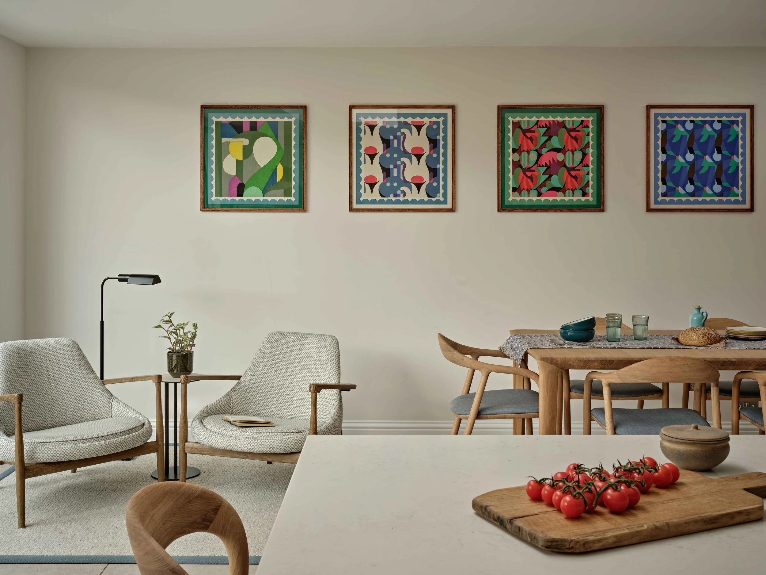 Interior of a modern dining and living room with four colorful framed abstract art pieces on the white wall, a wooden dining table with bowls and a loaf of bread, a white table with tomatoes on a wooden board, white armchairs, a wooden dining chair, 