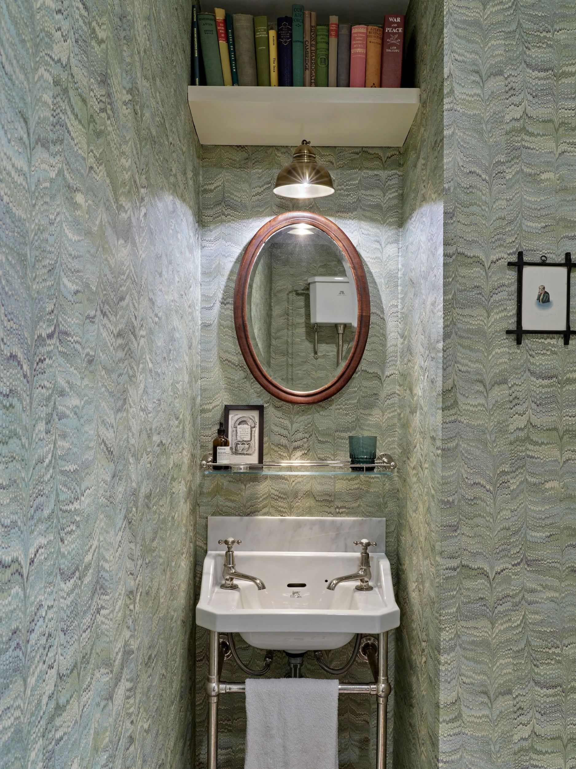 A small vintage-style bathroom with green and blue patterned wallpaper, a white porcelain sink with chrome fixtures, a towel hanging below, a round mirror with a wooden frame, a wall-mounted shelf with vintage books and a lamp, and a small framed pic