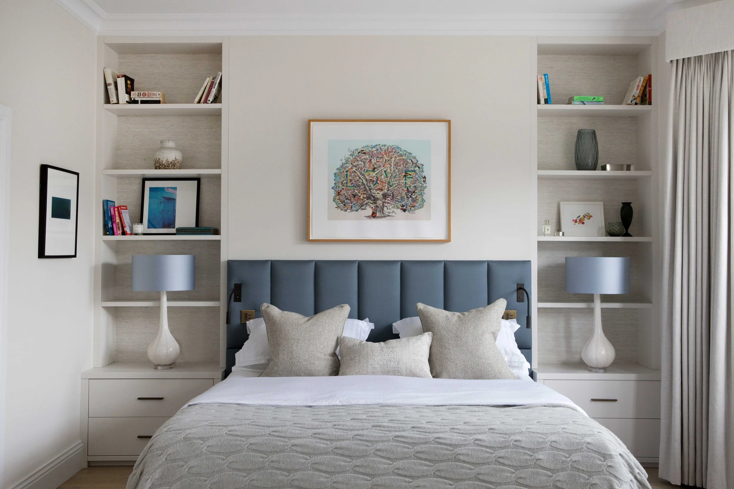 A bedroom with a bed, light grey headboard, decorative pillows, two white nightstands with matching lamps, built-in shelves on either side of the headboard holding books, framed pictures, and decorative objects, a framed artwork with a tree and the w