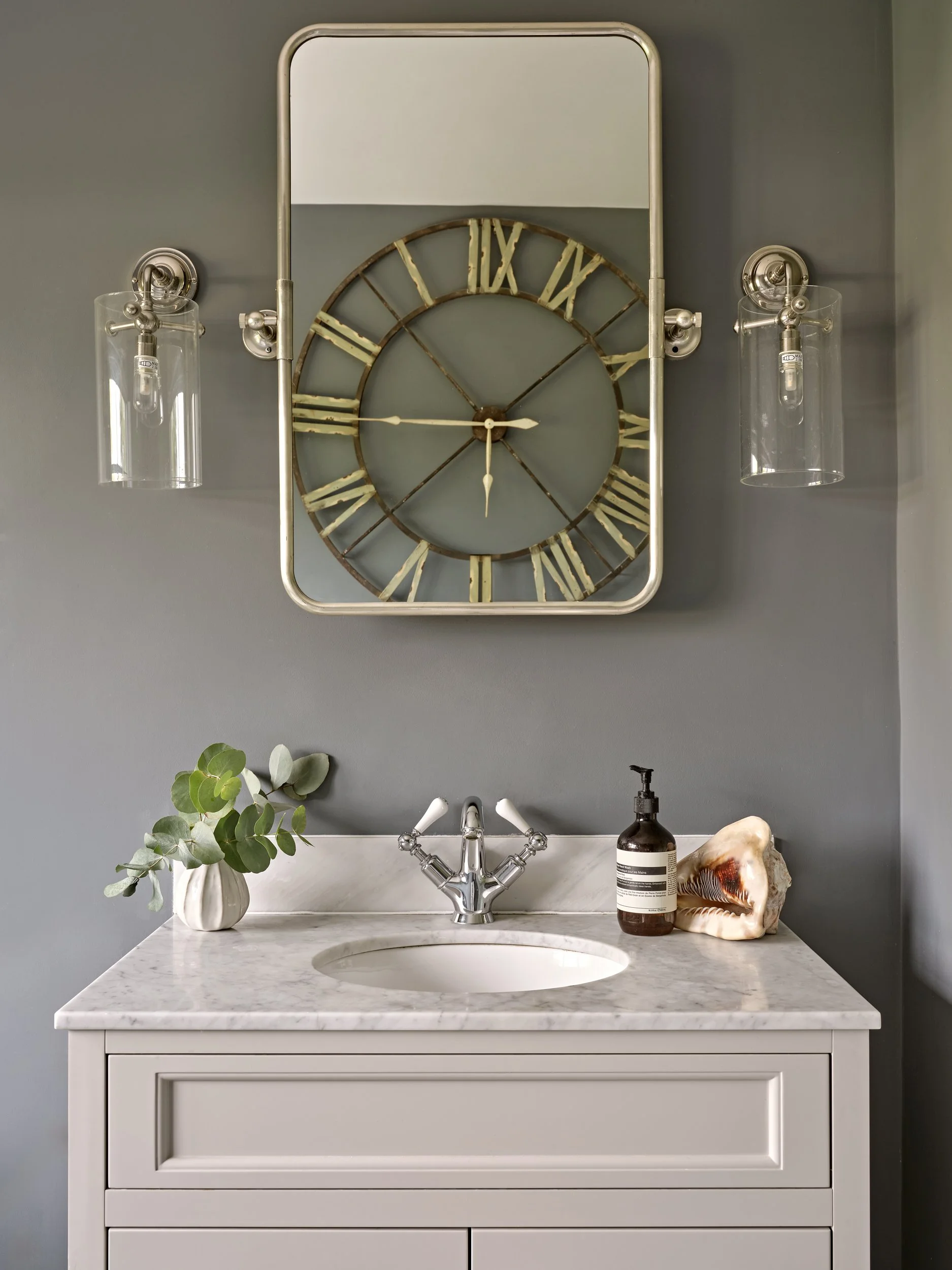 Bathroom vanity with marble countertop, white cabinet, and a round sink. On the countertop, there's a green leafy plant in a white vase, a brown bottle with a pump dispenser, and a large shell or coral piece. Behind the sink is a silver faucet. Above